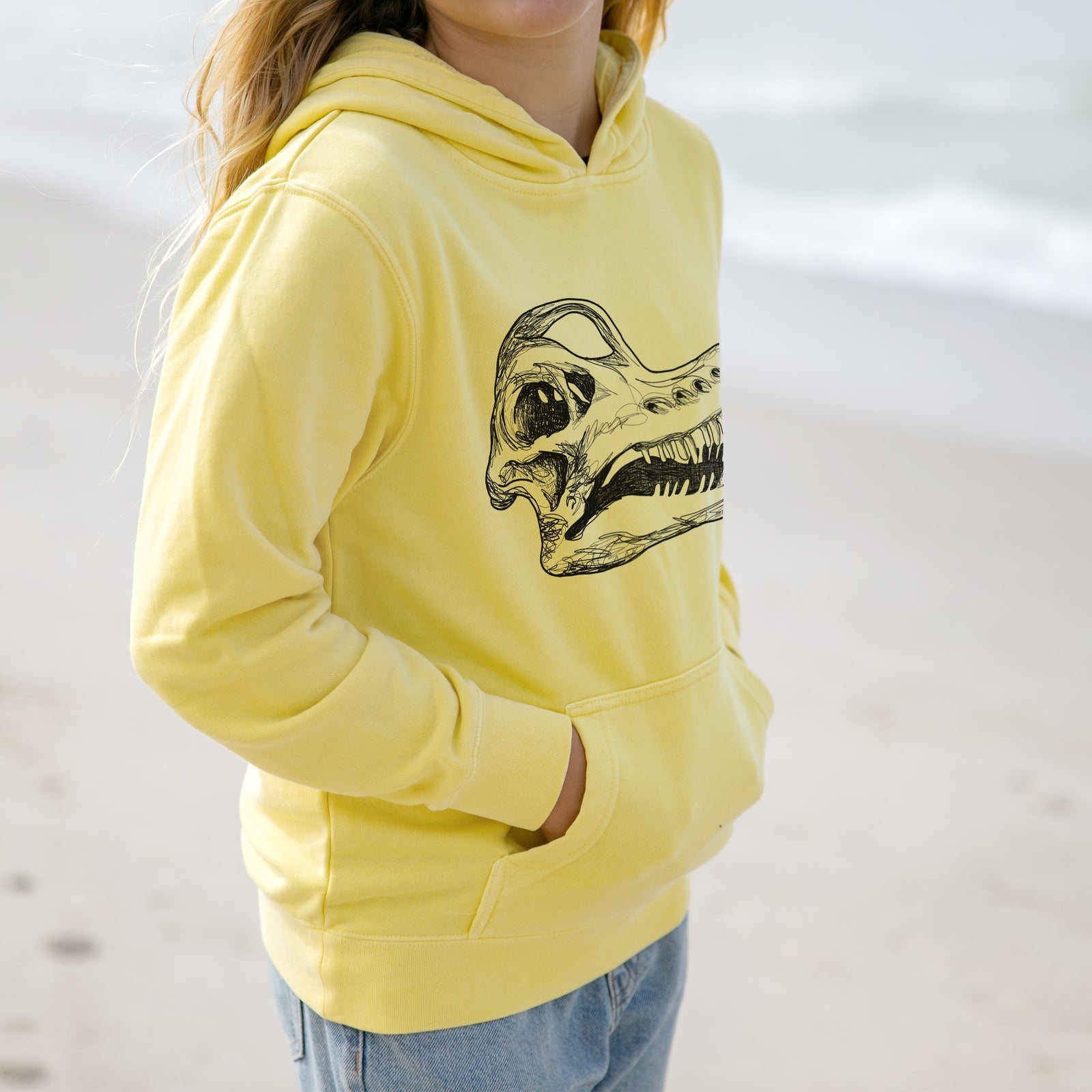 Boy wearing a youth pigment dyed hoodie printed with a detailed black ink illustration of a Brachiosaurus skull.