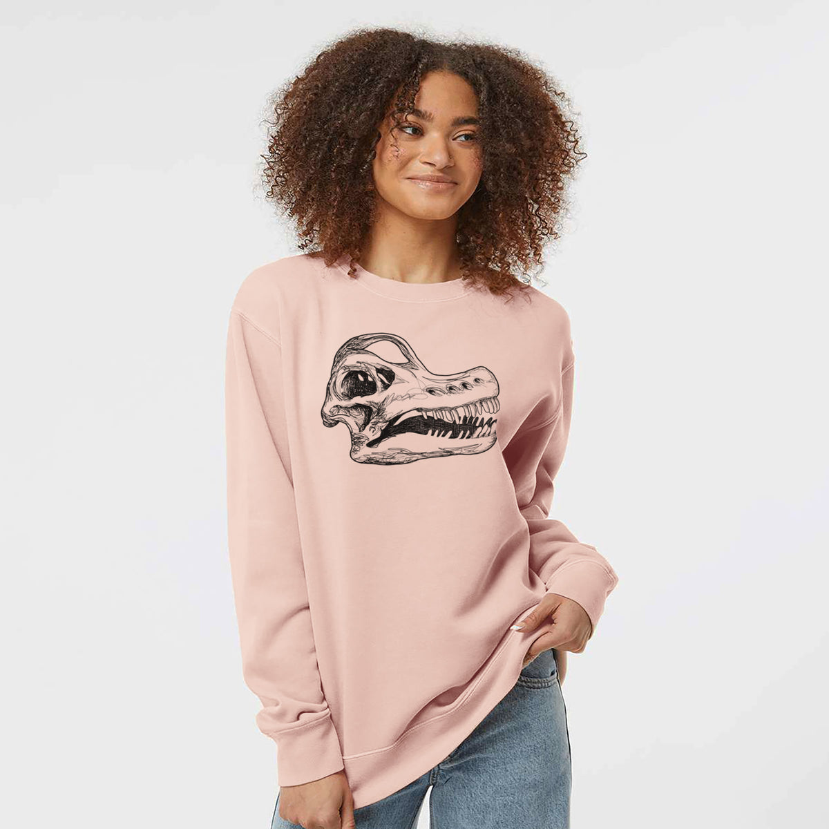 Model wearing a dusty pink pigment dyed crewneck sweatshirt printed with a detailed black ink illustration of a Brachiosaurus skull.