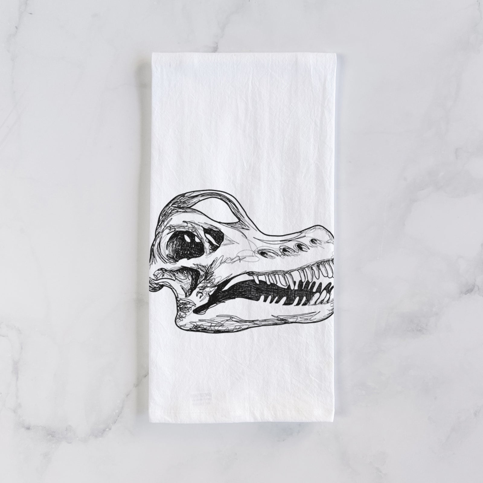 White cotton tea towel printed with a detailed black ink illustration of a Brachiosaurus skull, on a marble counter beside a round wooden cutting board and greenery.