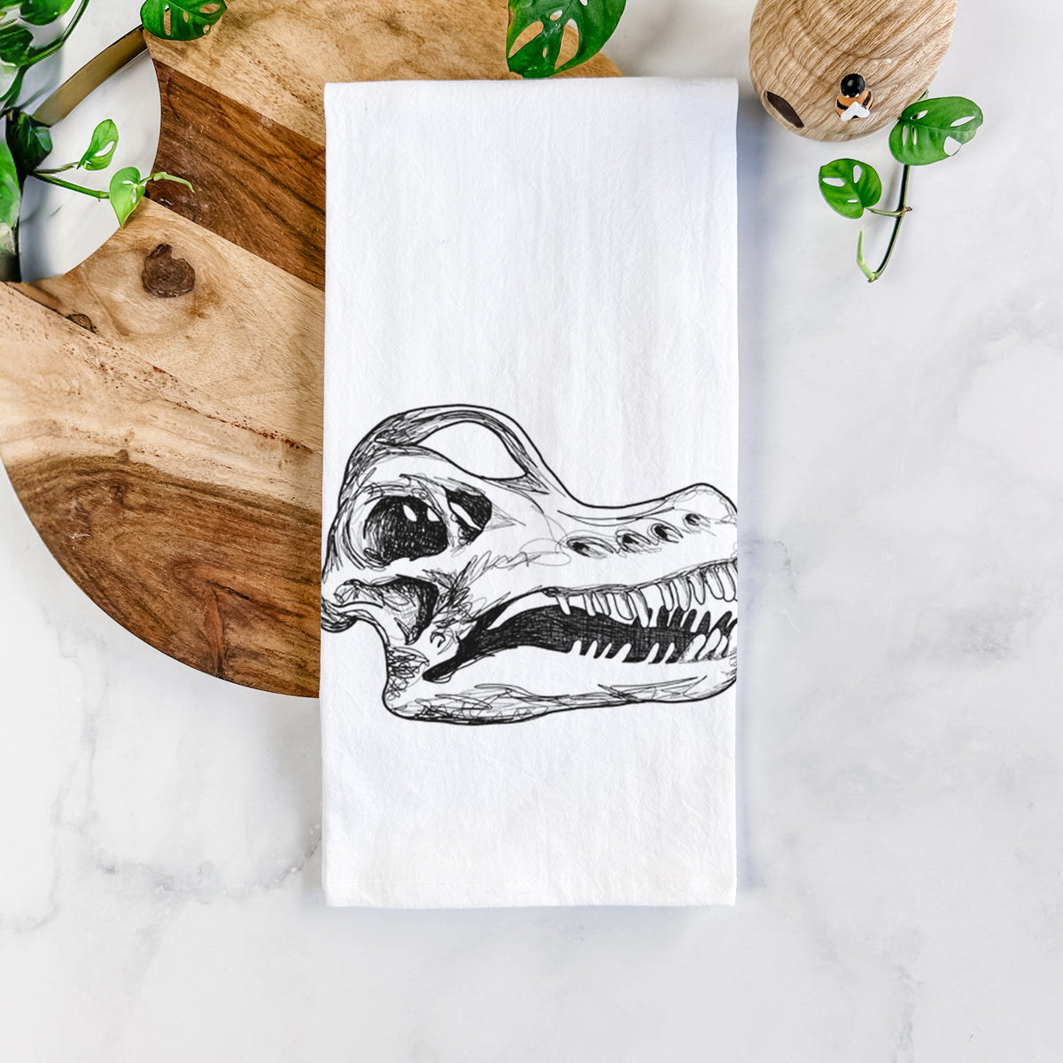 White cotton tea towel printed with a detailed black ink illustration of a Brachiosaurus skull, on a marble counter beside a round wooden cutting board and greenery.