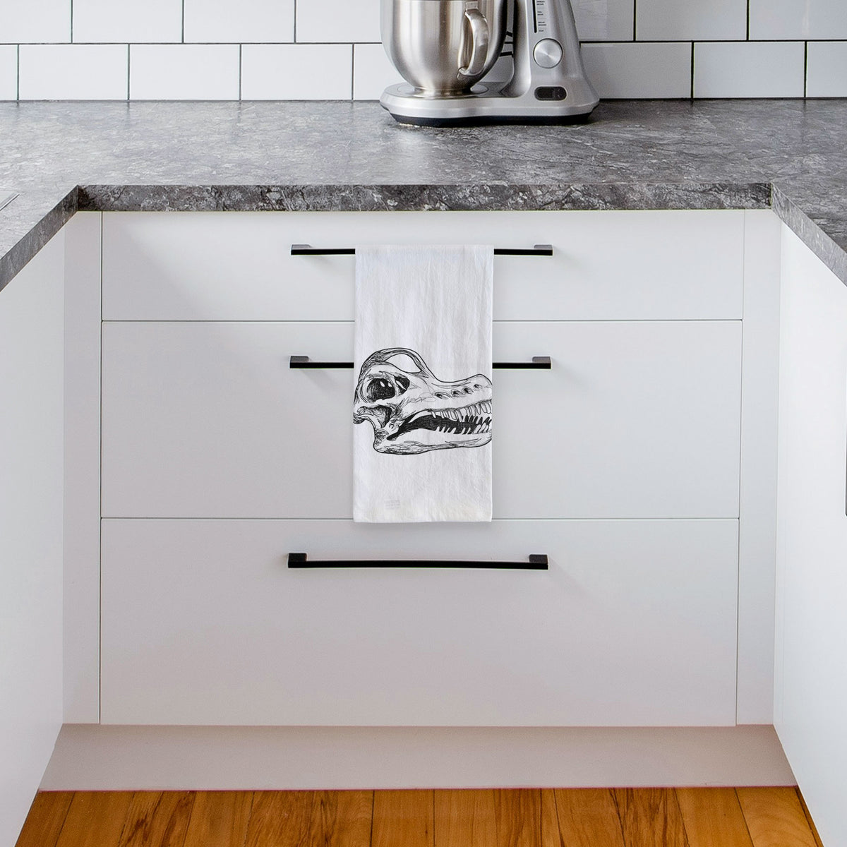 White tea towel printed with a detailed black ink illustration of a Brachiosaurus skull, hanging from a drawer handle in a bright white kitchen.