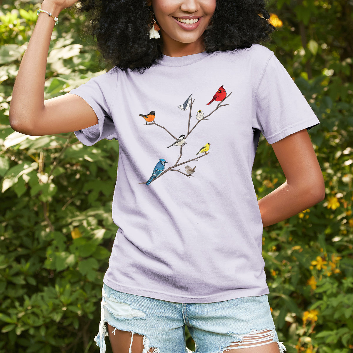 Woman wearing a Comfort Colors t-shirt printed with a full-color illustration of backyard birds, including a cardinal, blue jay, goldfinch and robin, perched on bare branches, outdoors.
