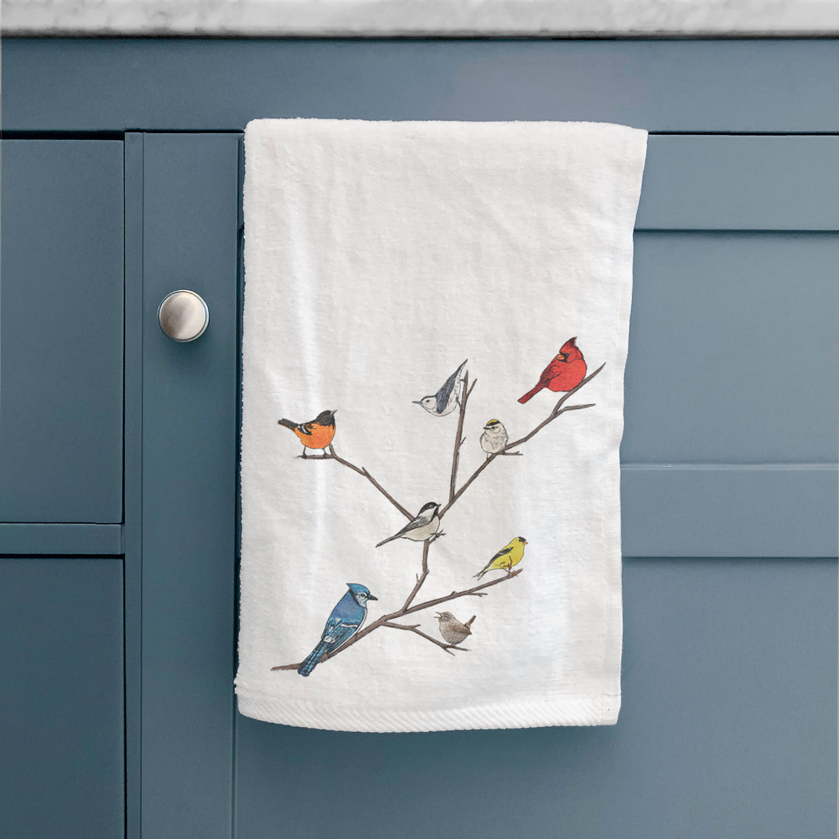White hand towel printed with a full-color illustration of backyard birds, including a cardinal, blue jay, goldfinch and robin, perched on bare branches, hanging on a cabinet door.