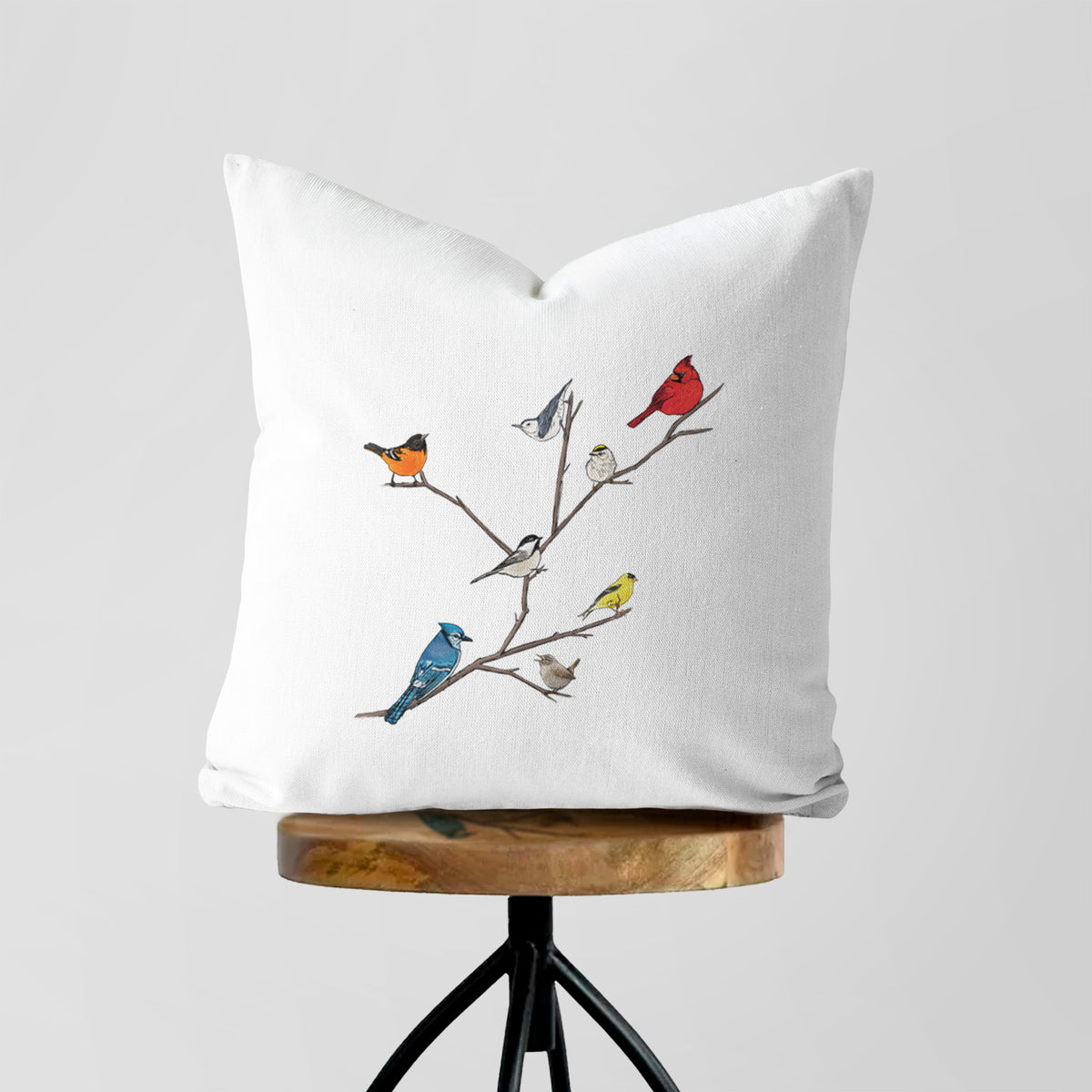 White pillow cover printed with a full-color illustration of backyard birds, including a cardinal, blue jay, goldfinch and robin, perched on bare branches, on a wooden stool.