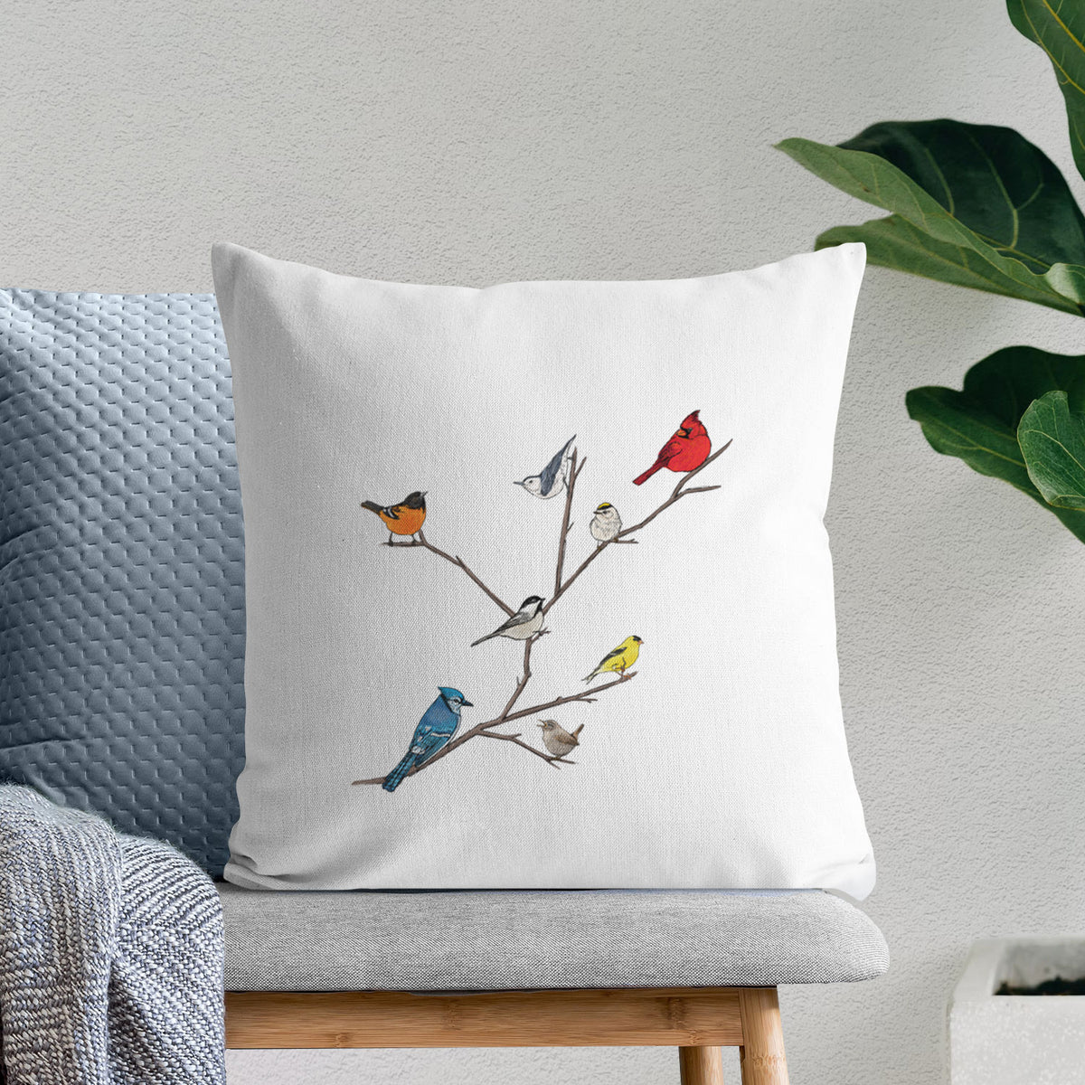 White pillow cover printed with a full-color illustration of backyard birds, including a cardinal, blue jay, goldfinch and robin, perched on bare branches, on a bench beside plants.