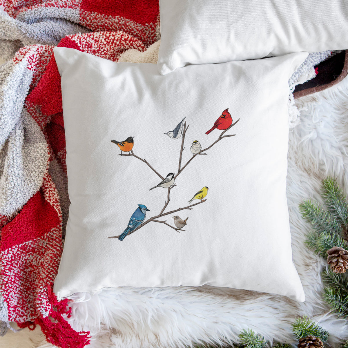 White pillow cover printed with a full-color illustration of backyard birds, including a cardinal, blue jay, goldfinch and robin, perched on bare branches, on a woven chair.