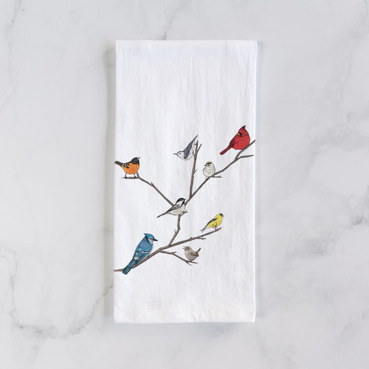 White cotton tea towel laid flat on marble, printed with a full-color illustration of backyard birds, including a cardinal, blue jay, goldfinch and robin, perched on bare branches.