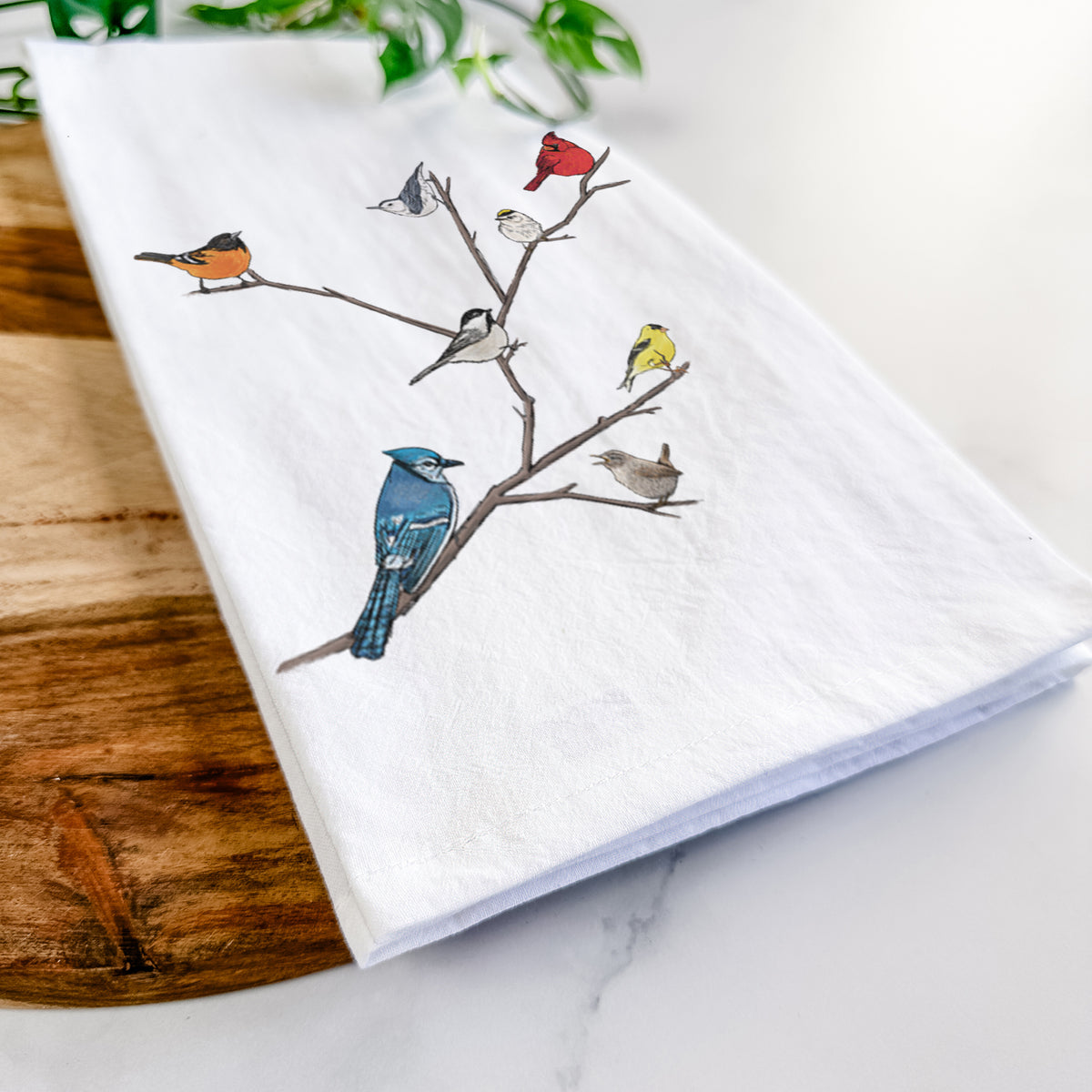 Close-up of a folded white tea towel on a wooden board, showing a full-color illustration of backyard birds, including a cardinal, blue jay, goldfinch and robin, perched on bare branches.