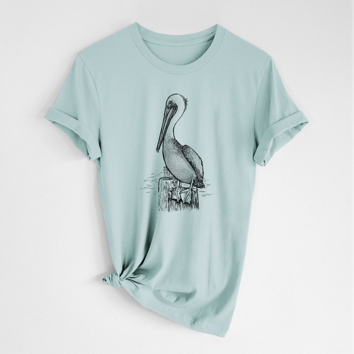 Dusty blue lightweight cotton t-shirt laid flat with the sleeves rolled, printed with a detailed black ink illustration of a brown pelican perched on a wooden piling.