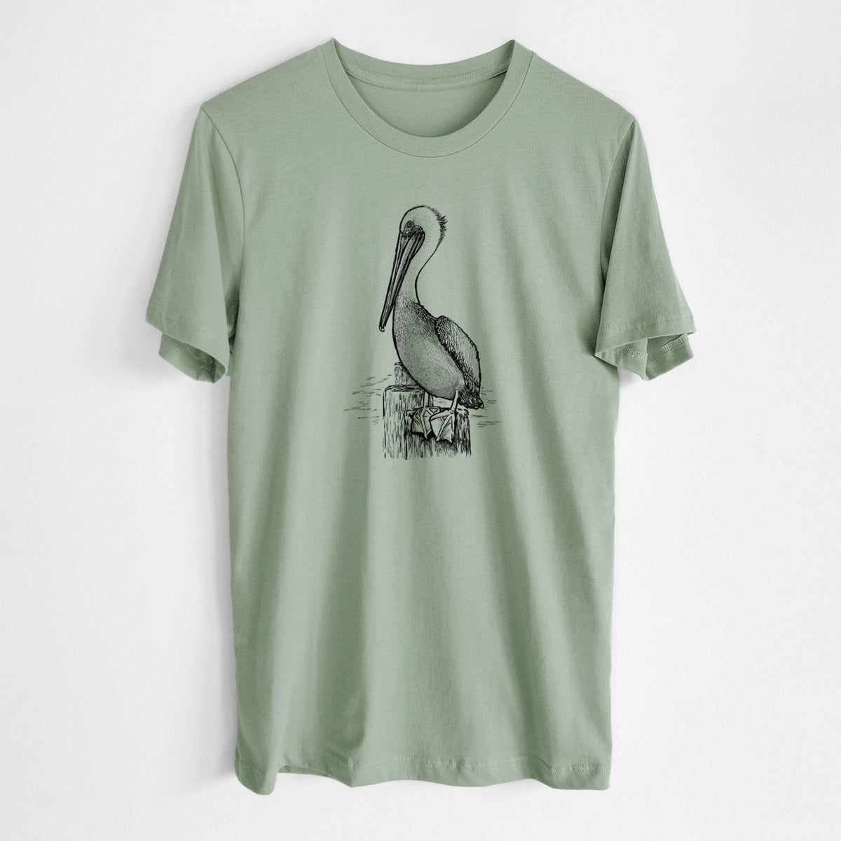 Sage green lightweight cotton t-shirt laid flat, printed with a detailed black ink illustration of a brown pelican perched on a wooden piling.
