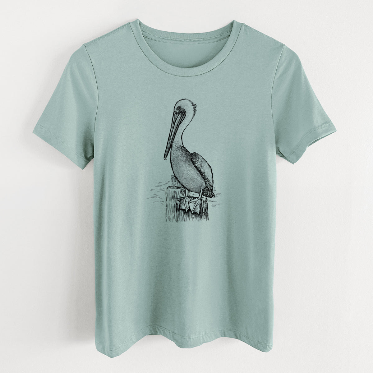 Dusty blue women's relaxed fit cotton t-shirt laid flat, printed with a detailed black ink illustration of a brown pelican perched on a wooden piling.