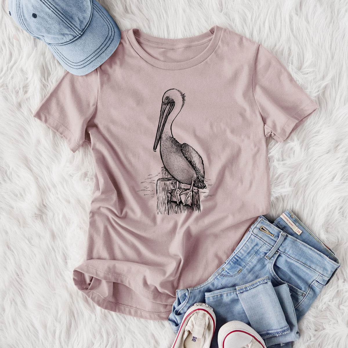 Women's relaxed fit cotton t-shirt printed with a detailed black ink illustration of a brown pelican perched on a wooden piling, styled flat on a white faux fur rug with jeans.