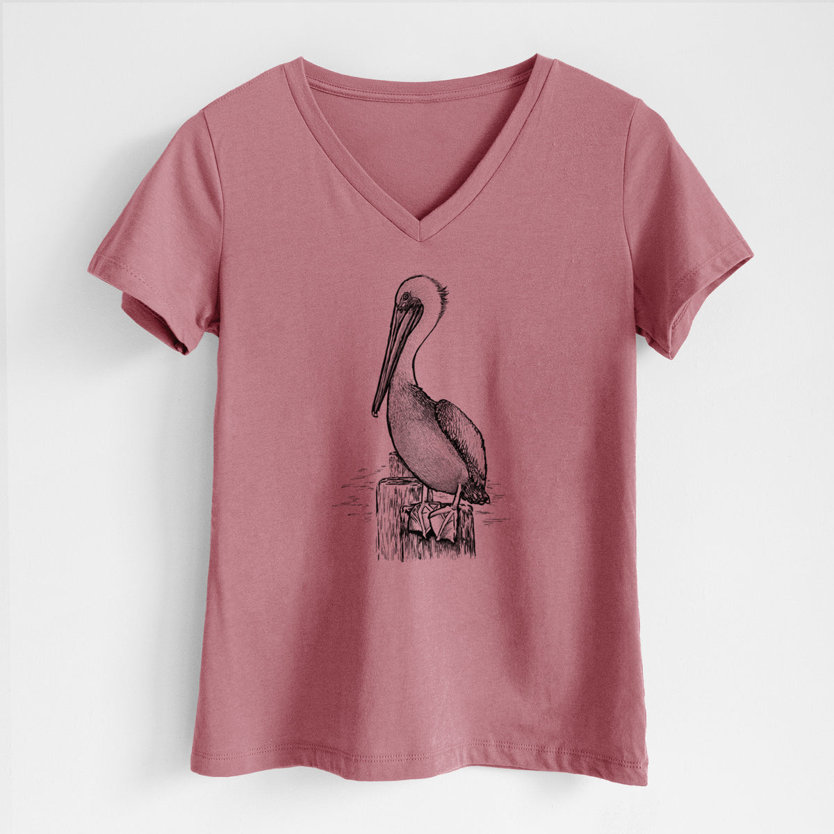 Mauve pink women's relaxed fit cotton V-neck t-shirt laid flat, printed with a detailed black ink illustration of a brown pelican perched on a wooden piling.