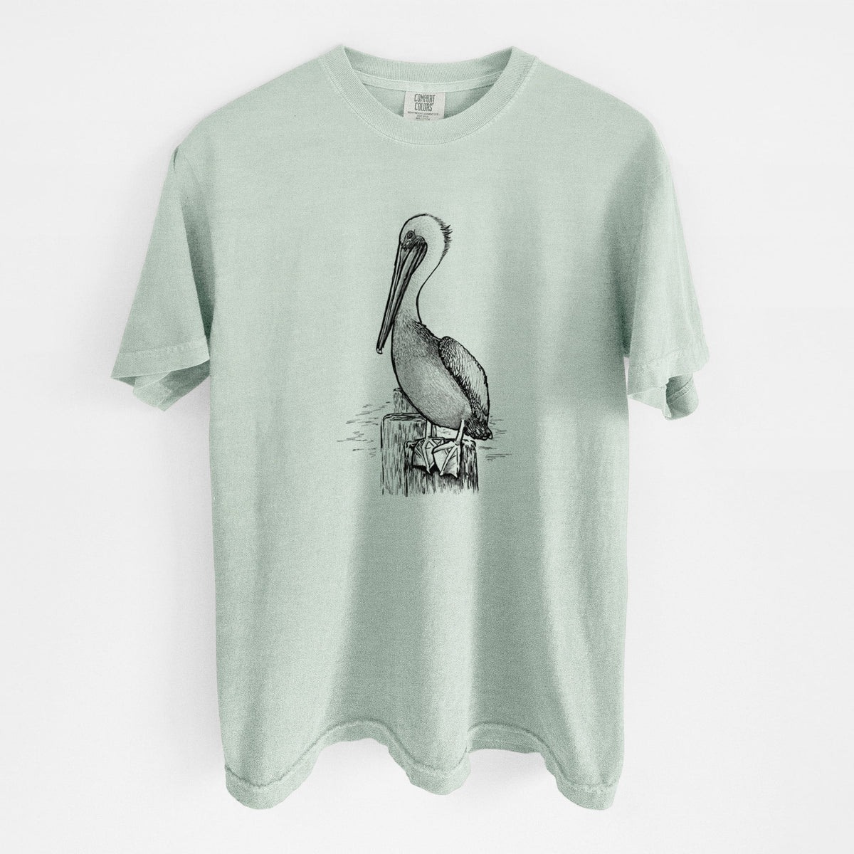 Bay green Comfort Colors t-shirt laid flat, printed with a detailed black ink illustration of a brown pelican perched on a wooden piling.