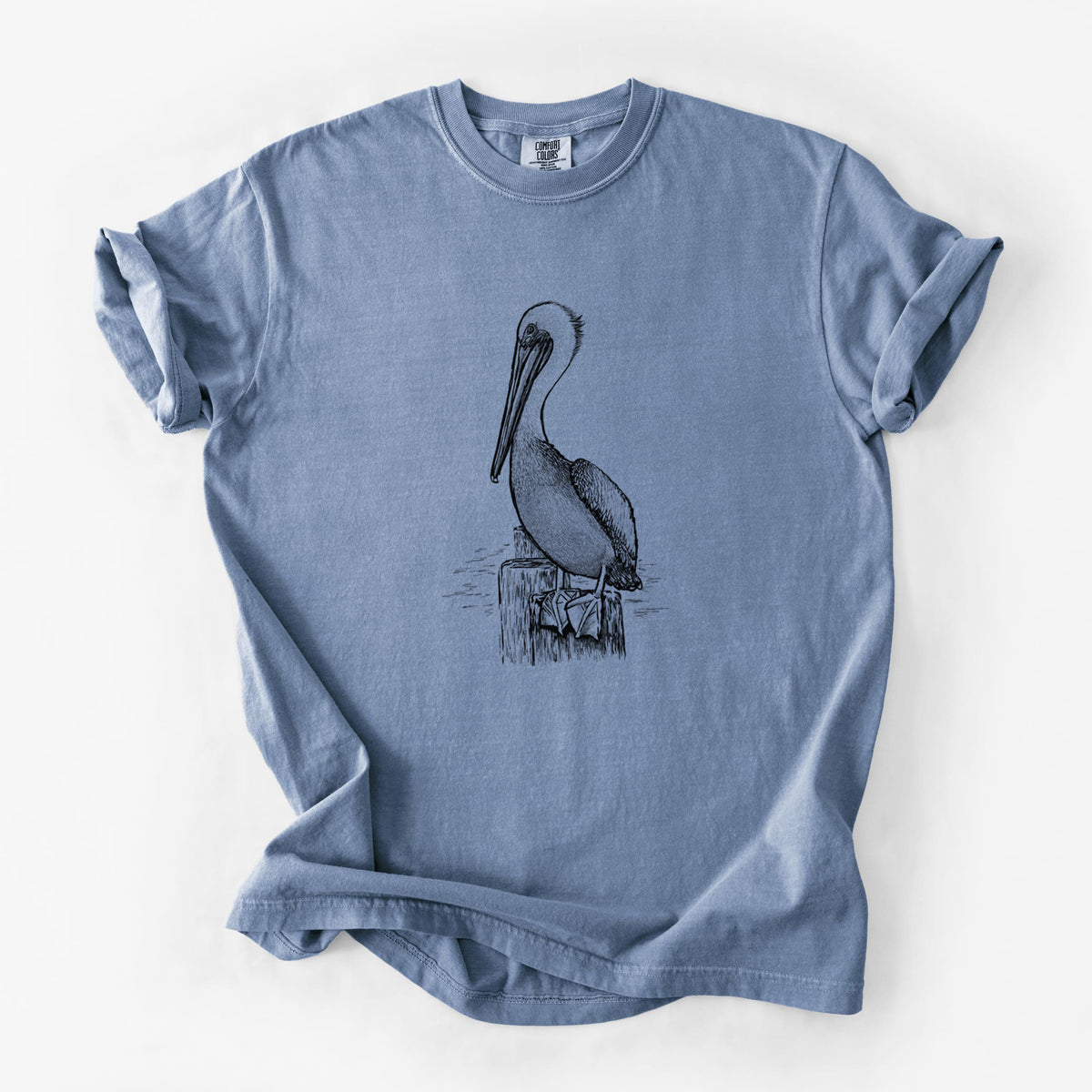 Blue jean Comfort Colors t-shirt laid flat, printed with a detailed black ink illustration of a brown pelican perched on a wooden piling.