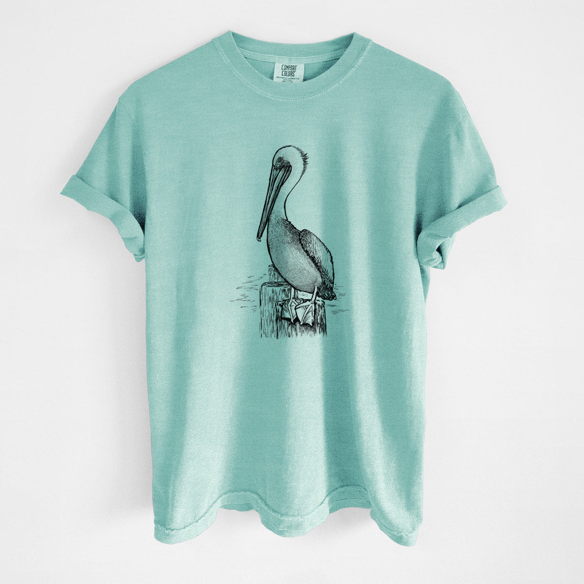 Chalky mint Comfort Colors t-shirt laid flat, printed with a detailed black ink illustration of a brown pelican perched on a wooden piling.