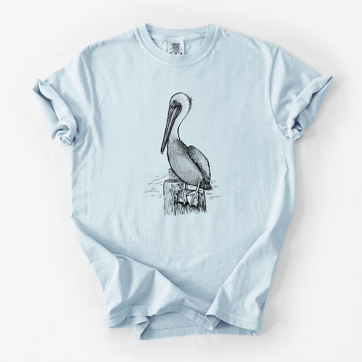 Chambray blue Comfort Colors t-shirt laid flat, printed with a detailed black ink illustration of a brown pelican perched on a wooden piling.