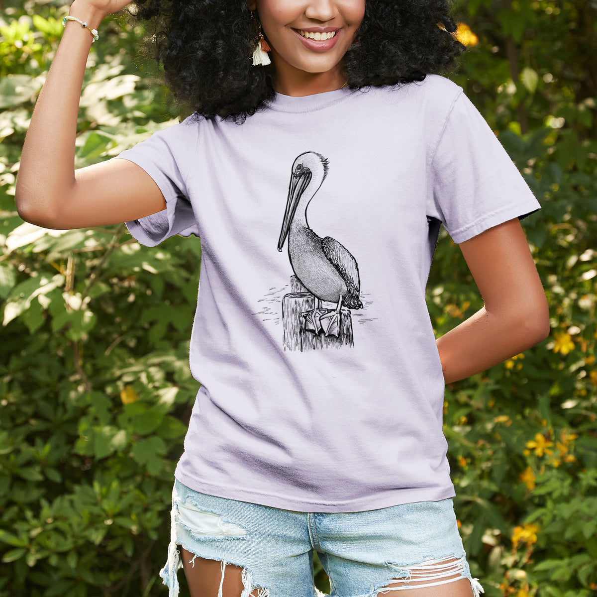 Woman wearing a Comfort Colors t-shirt printed with a detailed black ink illustration of a brown pelican perched on a wooden piling, outdoors.