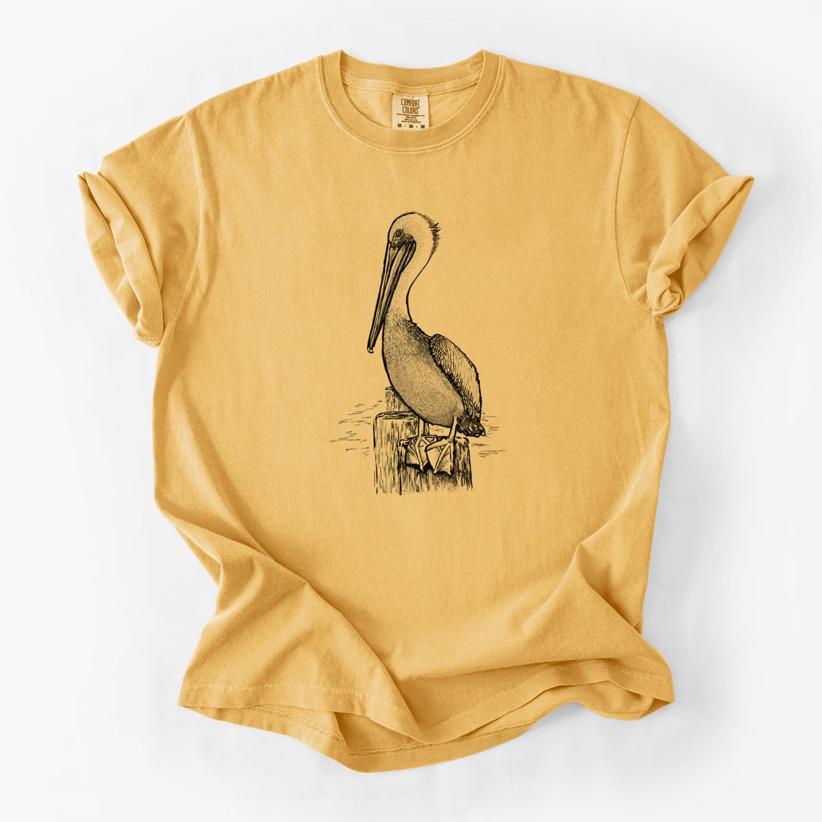 Golden wheat yellow Comfort Colors t-shirt laid flat, printed with a detailed black ink illustration of a brown pelican perched on a wooden piling.