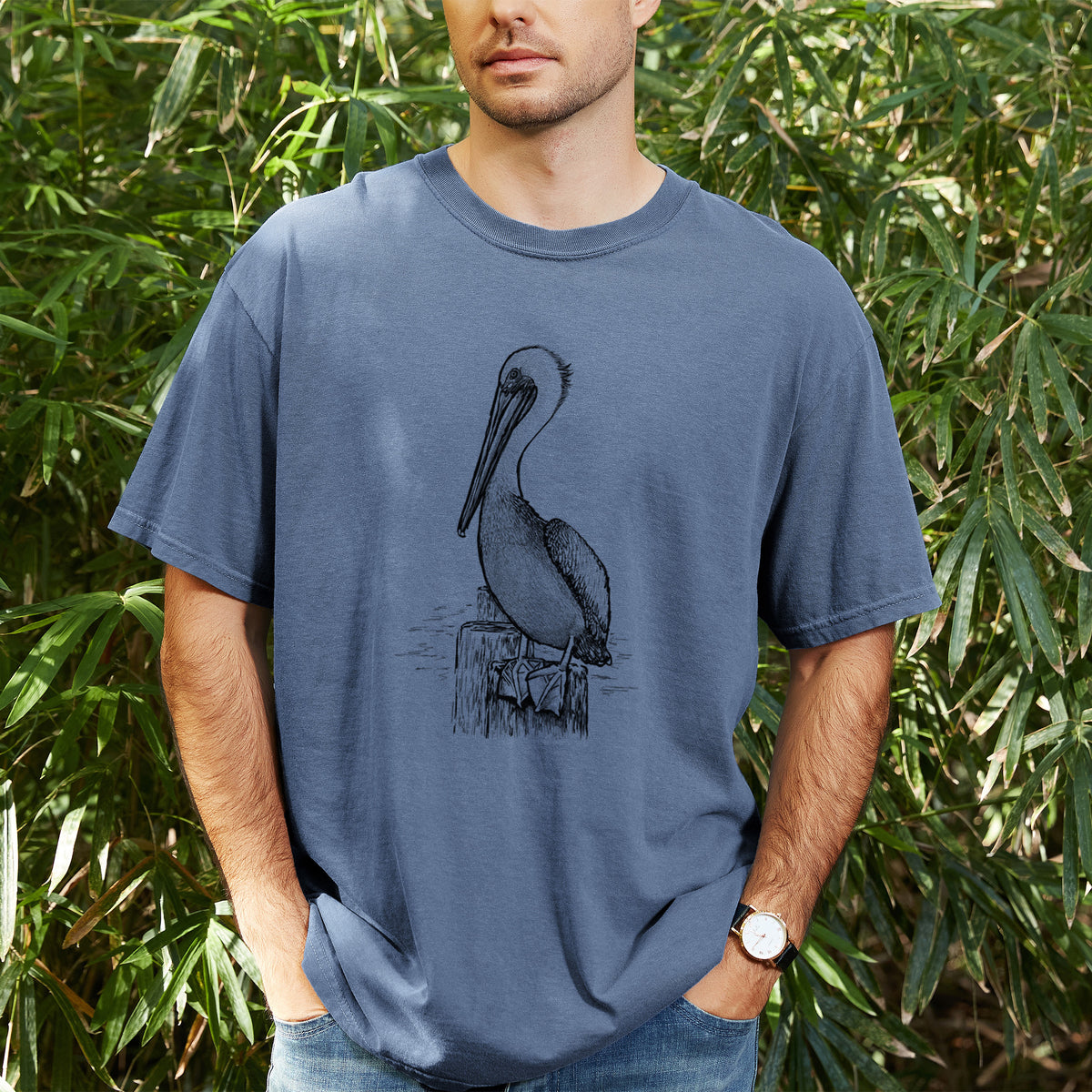 Man wearing a Comfort Colors t-shirt printed with a detailed black ink illustration of a brown pelican perched on a wooden piling, outdoors.