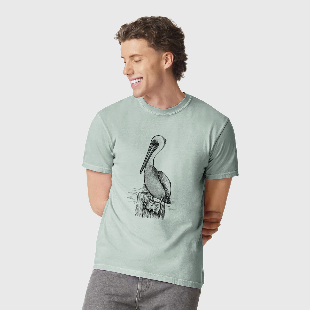Man wearing a Comfort Colors t-shirt printed with a detailed black ink illustration of a brown pelican perched on a wooden piling.
