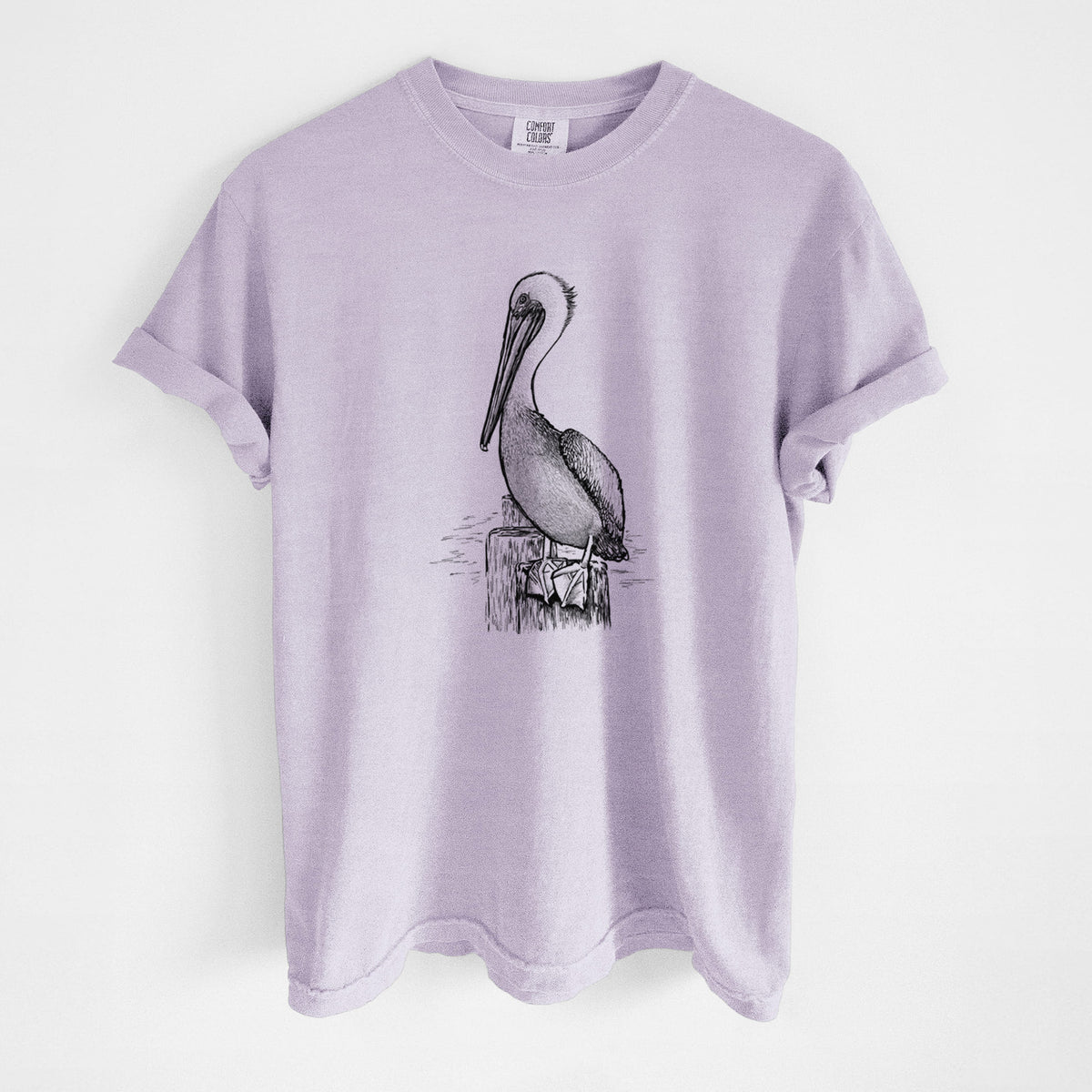 Orchid purple Comfort Colors t-shirt laid flat, printed with a detailed black ink illustration of a brown pelican perched on a wooden piling.