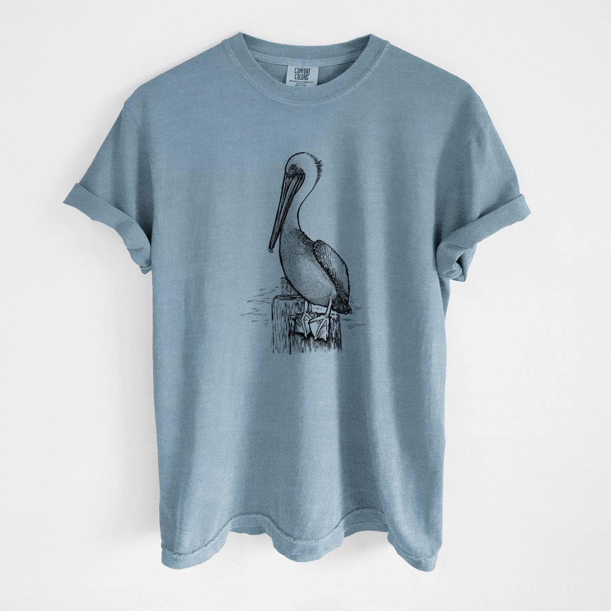 Steel blue Comfort Colors t-shirt laid flat, printed with a detailed black ink illustration of a brown pelican perched on a wooden piling.