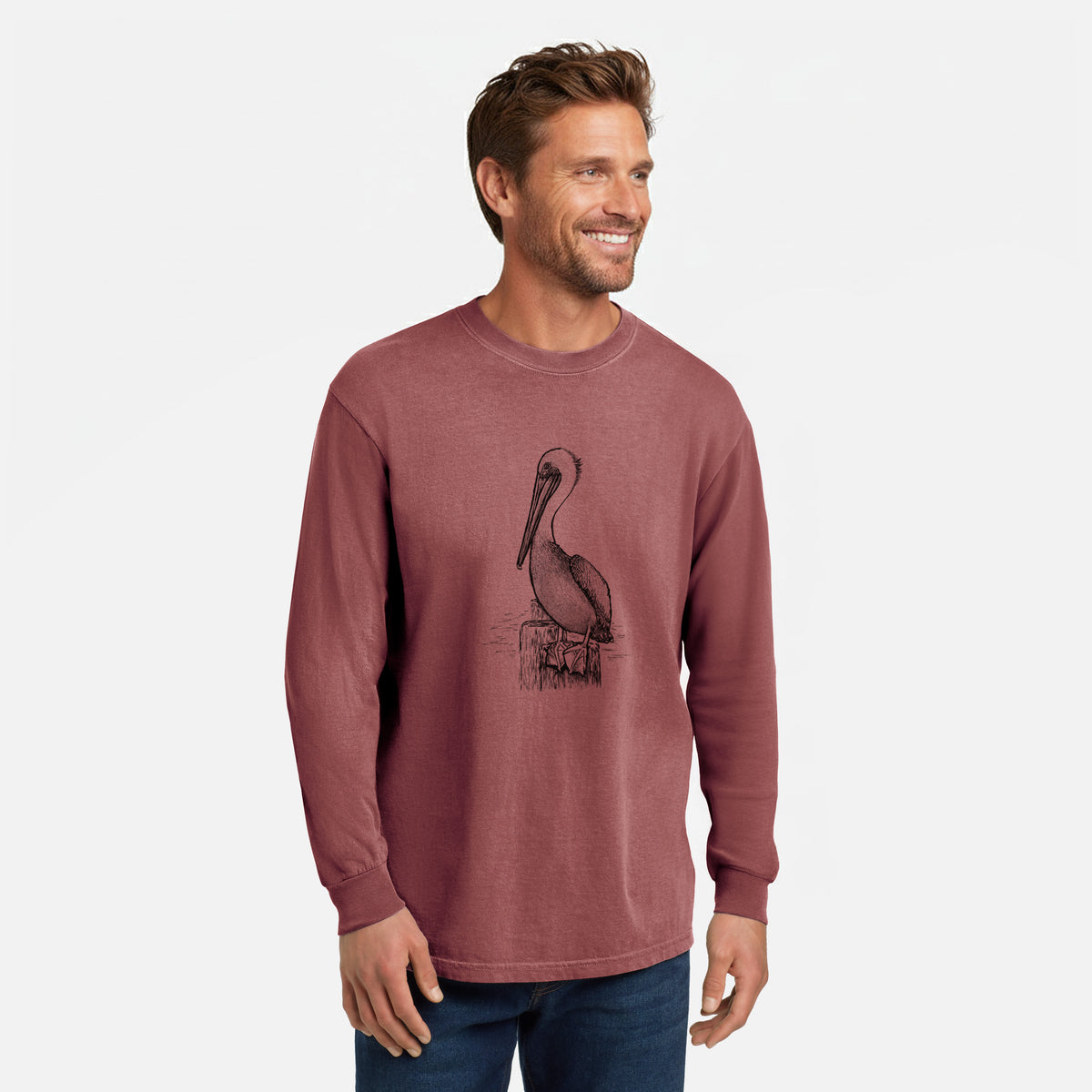 Brick red Comfort Colors long sleeve shirt printed with a detailed black ink illustration of a brown pelican perched on a wooden piling, shown in a lifestyle photo.
