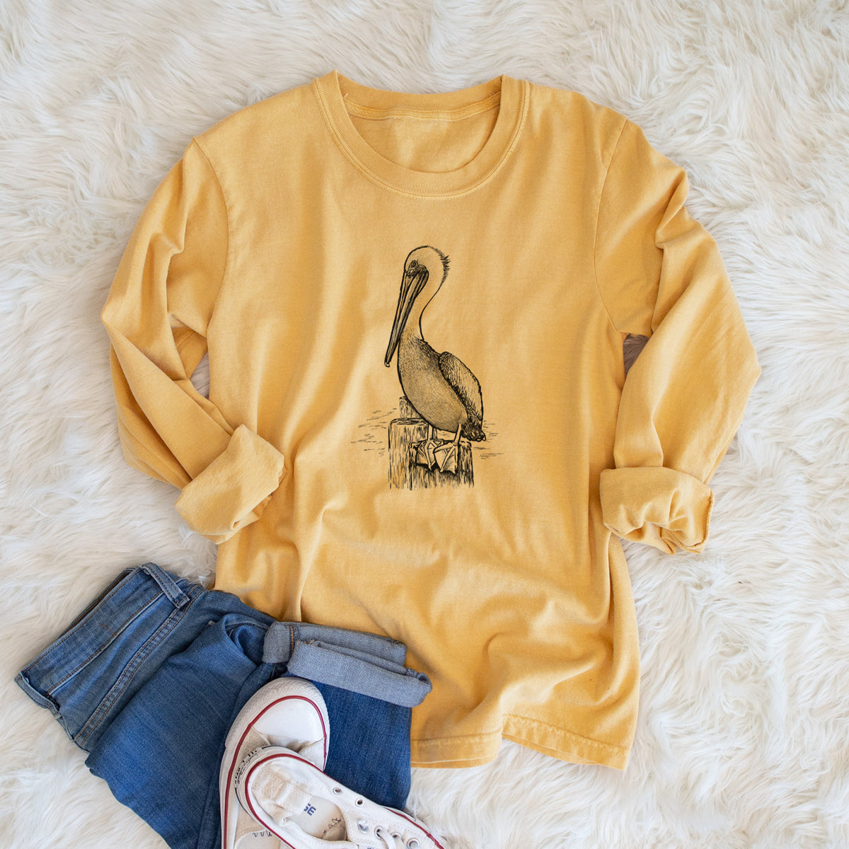 Golden wheat yellow Comfort Colors long sleeve shirt printed with a detailed black ink illustration of a brown pelican perched on a wooden piling, shown in a lifestyle photo.