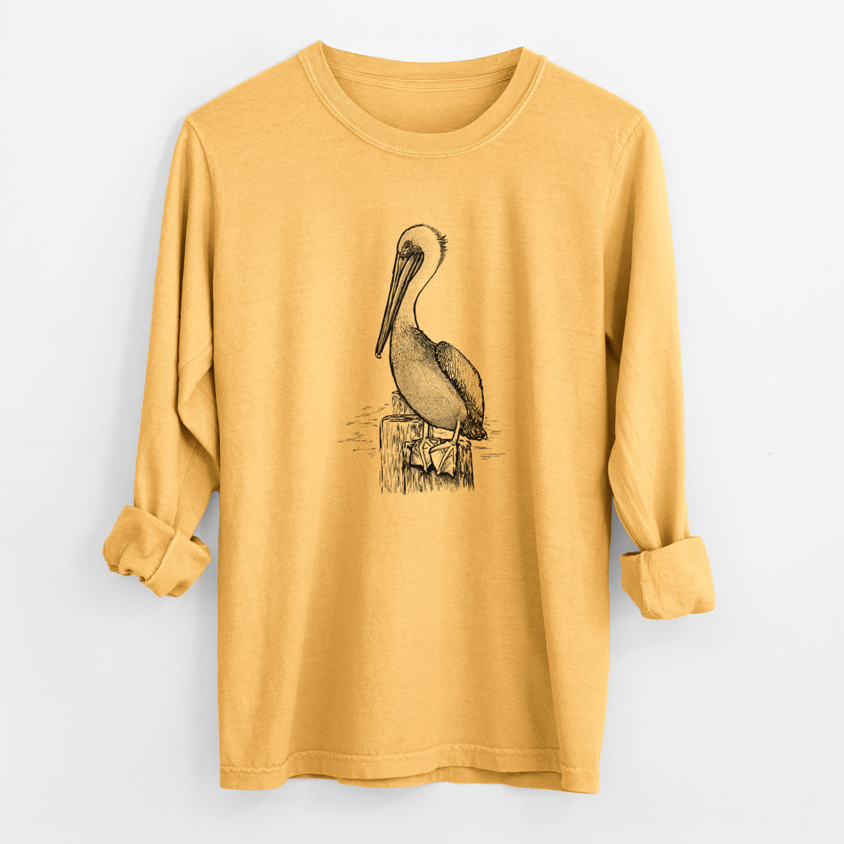 Golden wheat yellow Comfort Colors long sleeve shirt laid flat with the cuffs rolled, printed with a detailed black ink illustration of a brown pelican perched on a wooden piling.