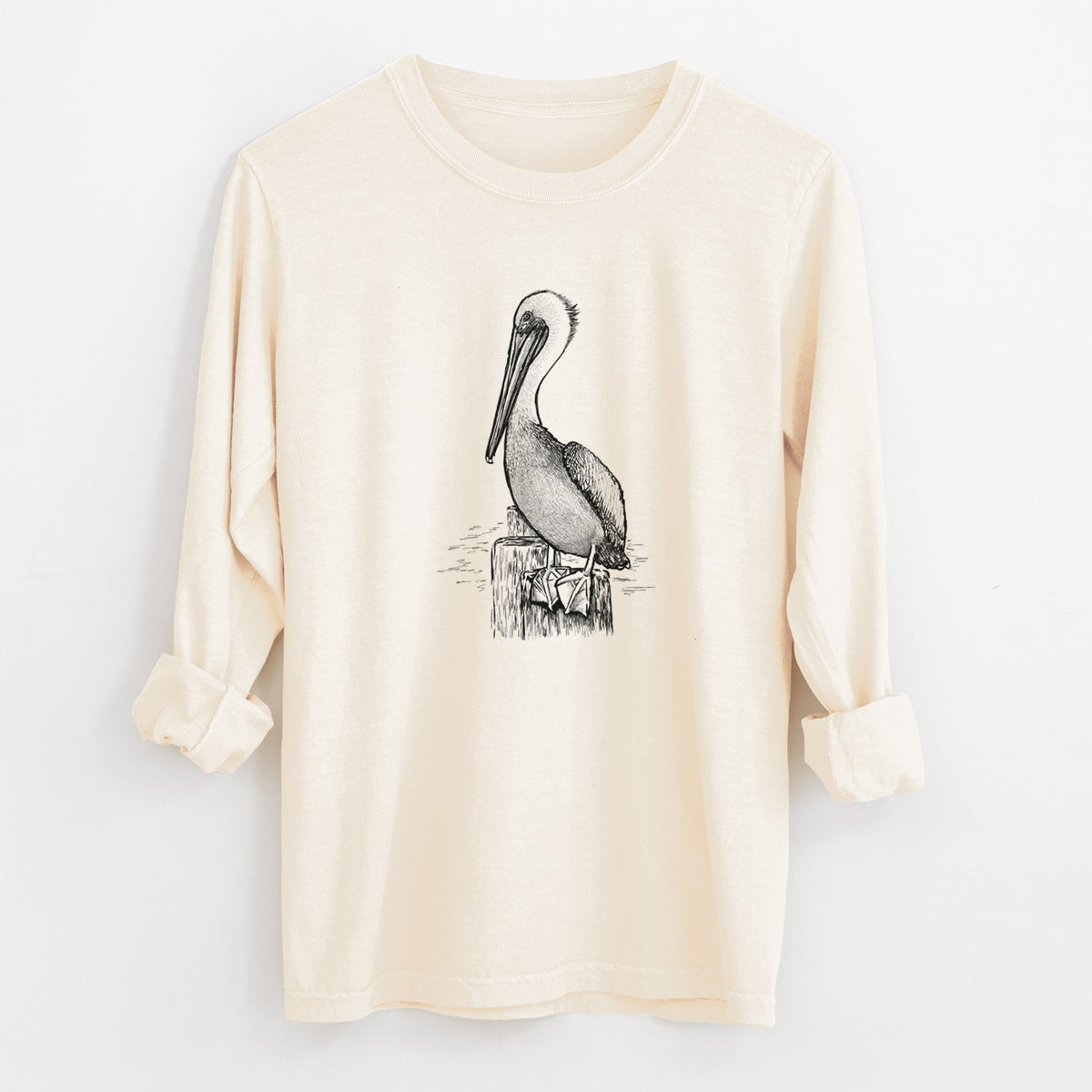 Ivory Comfort Colors long sleeve shirt laid flat with the cuffs rolled, printed with a detailed black ink illustration of a brown pelican perched on a wooden piling.