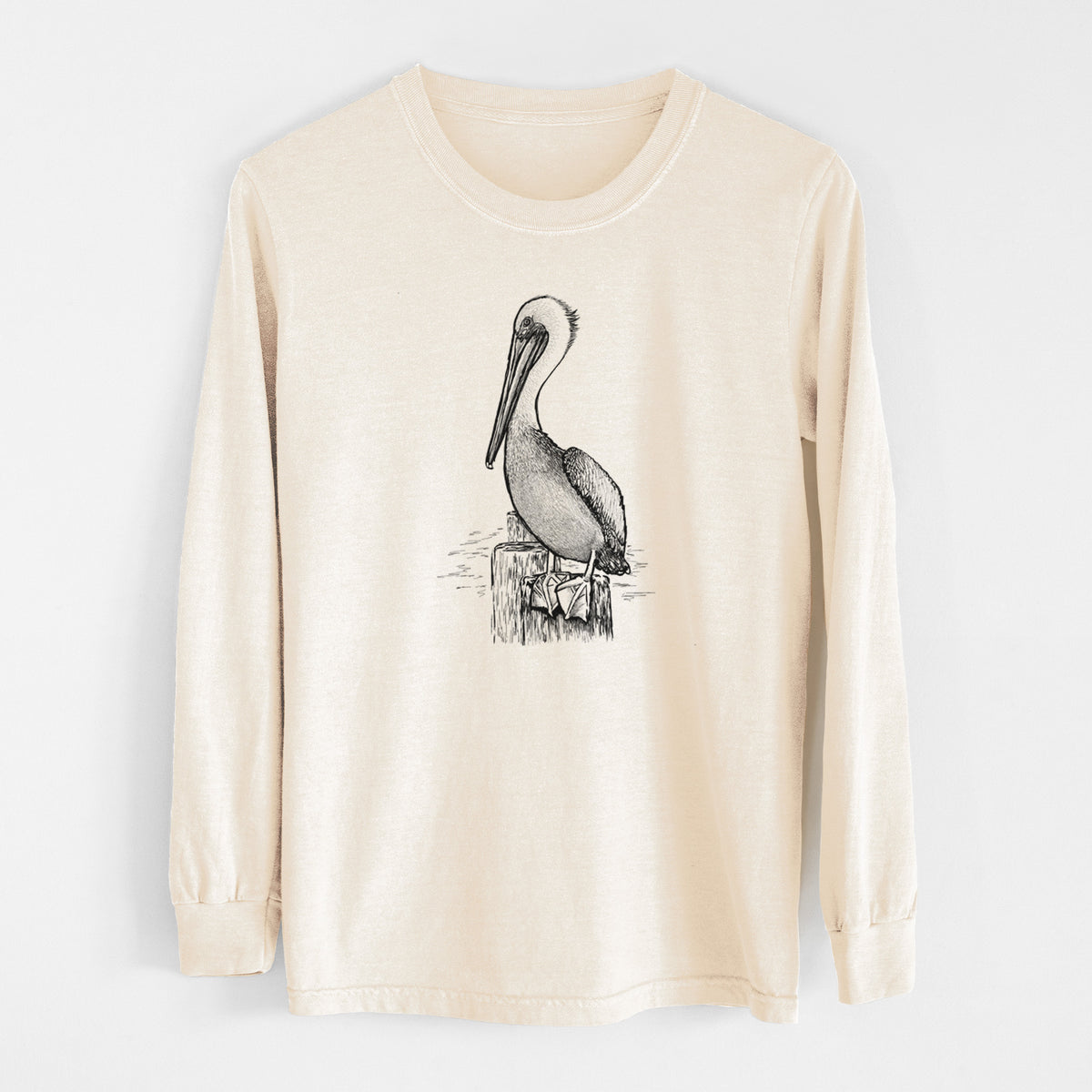 Ivory Comfort Colors long sleeve shirt laid flat, printed with a detailed black ink illustration of a brown pelican perched on a wooden piling.