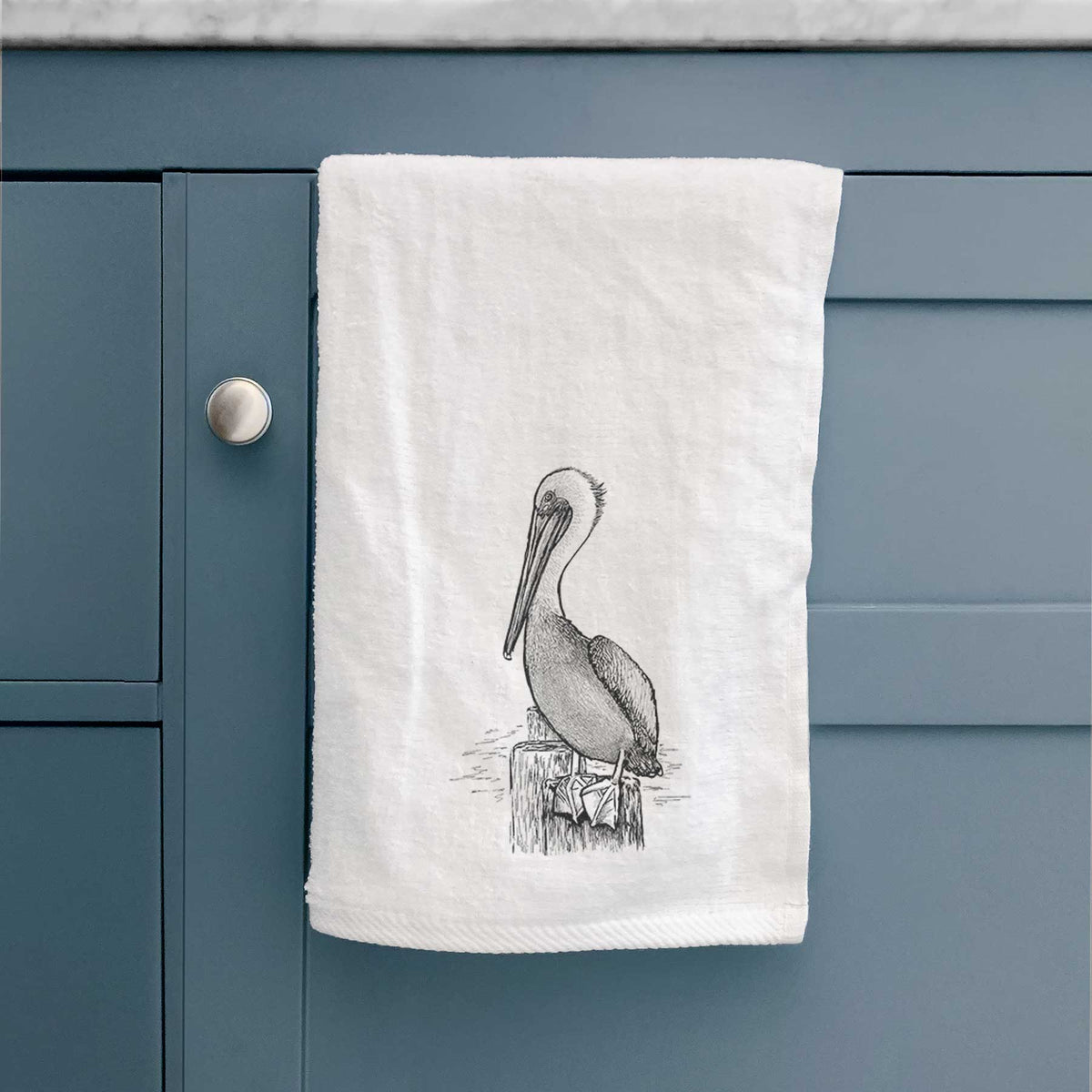 White hand towel printed with a detailed black ink illustration of a brown pelican perched on a wooden piling, hanging on a cabinet door.