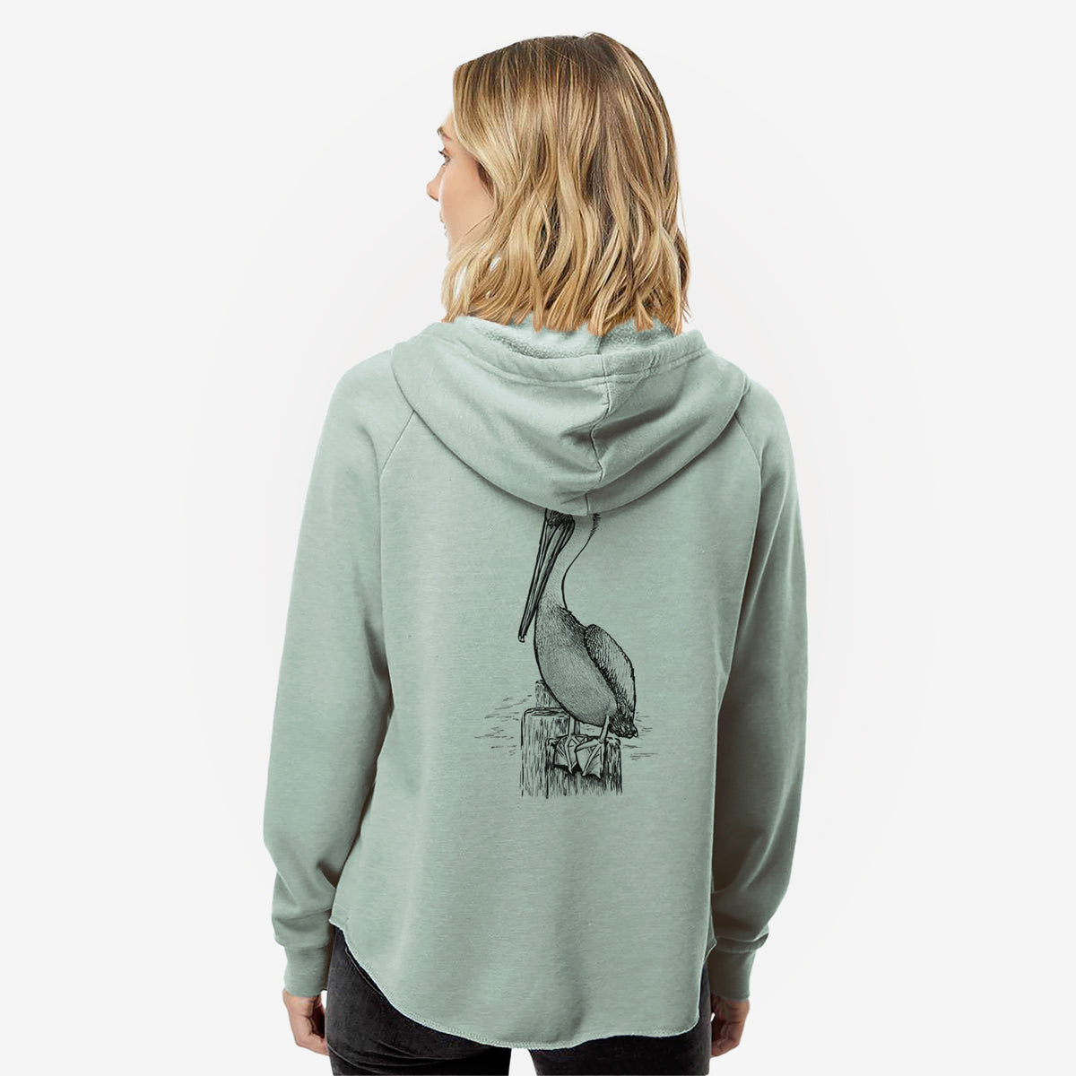 Woman wearing a sage green women's Cali Wave zip-up sweatshirt printed with a detailed black ink illustration of a brown pelican perched on a wooden piling, seen from behind.