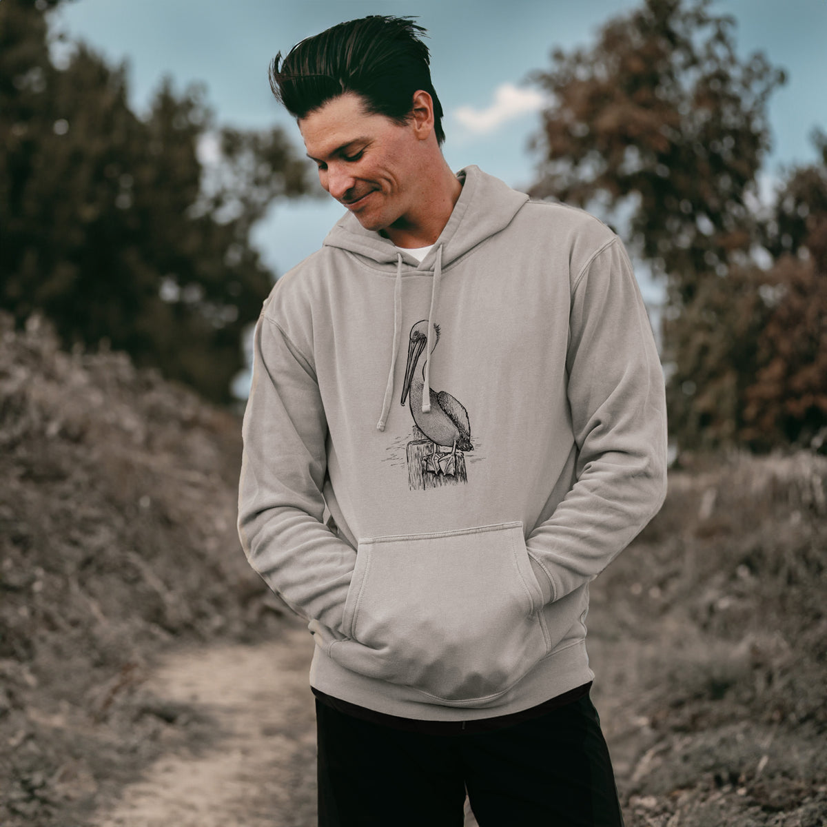 Man wearing a cement grey pigment dyed hoodie printed with a detailed black ink illustration of a brown pelican perched on a wooden piling.