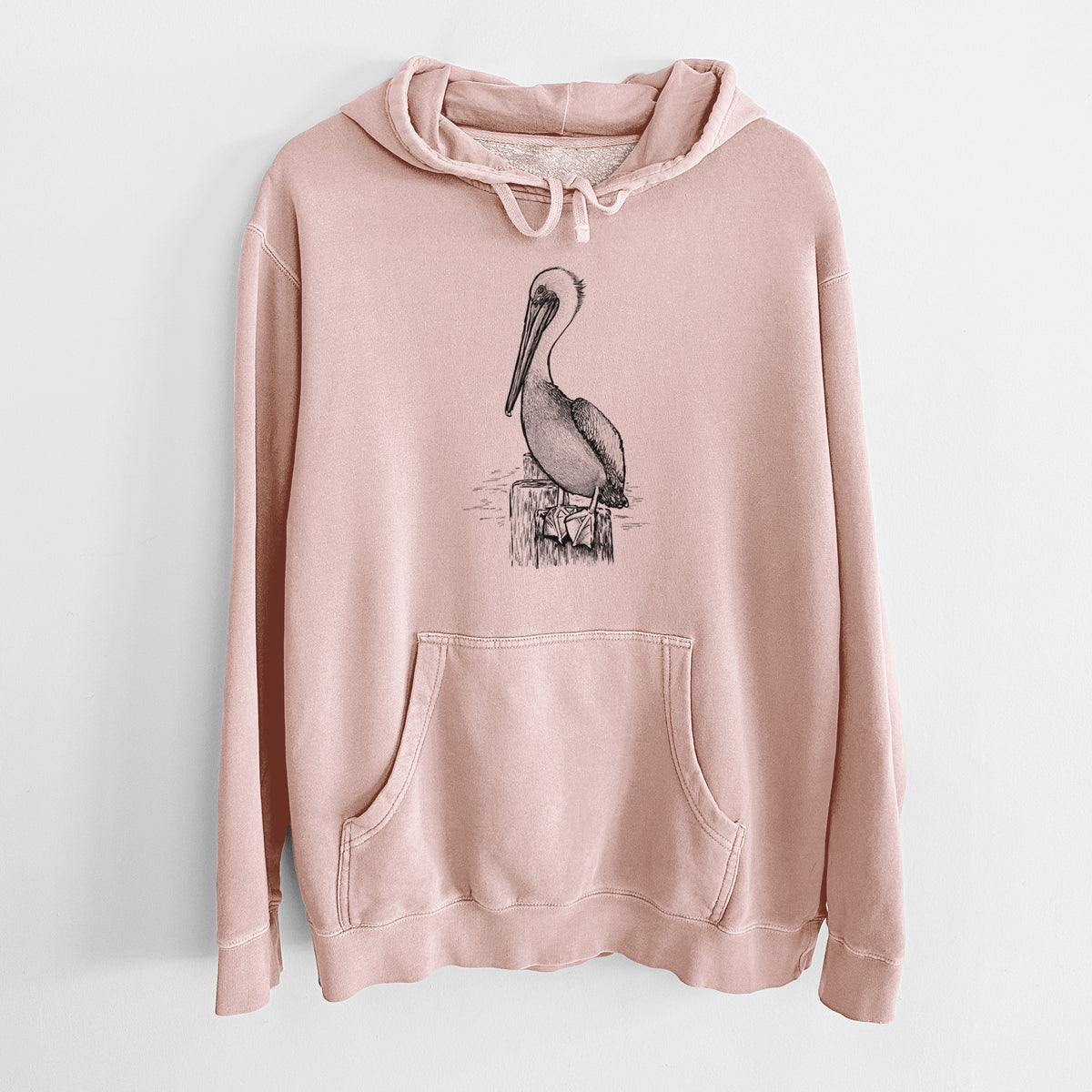 Dusty pink pigment dyed hoodie laid flat, printed with a detailed black ink illustration of a brown pelican perched on a wooden piling.