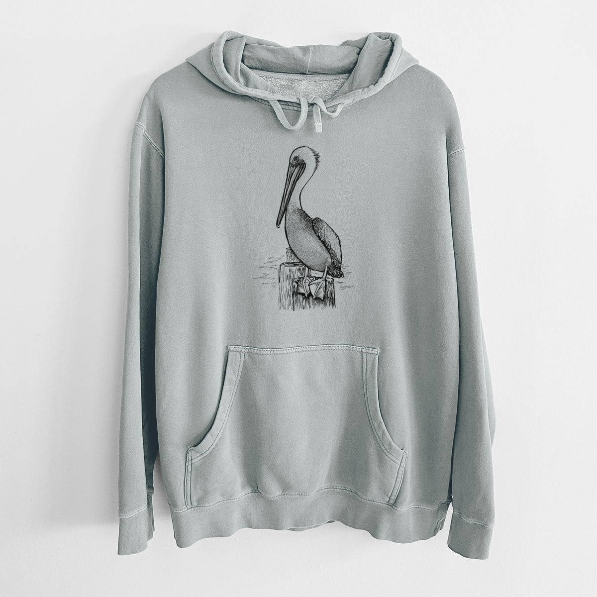 Sage green pigment dyed hoodie laid flat, printed with a detailed black ink illustration of a brown pelican perched on a wooden piling.