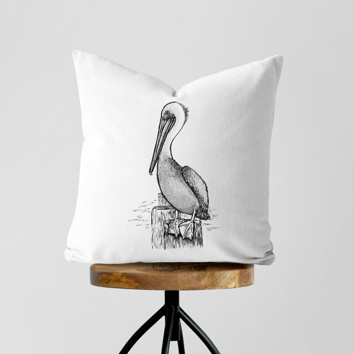 White pillow cover printed with a detailed black ink illustration of a brown pelican perched on a wooden piling, on a wooden stool.