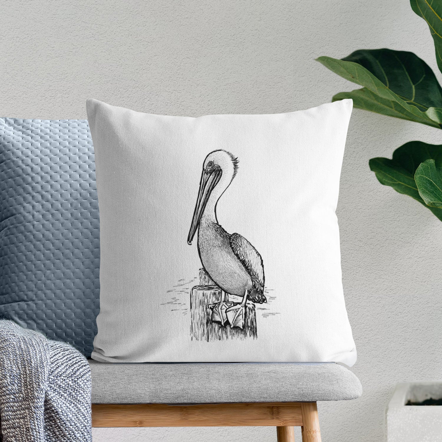 White pillow cover printed with a detailed black ink illustration of a brown pelican perched on a wooden piling, on a bench beside plants.