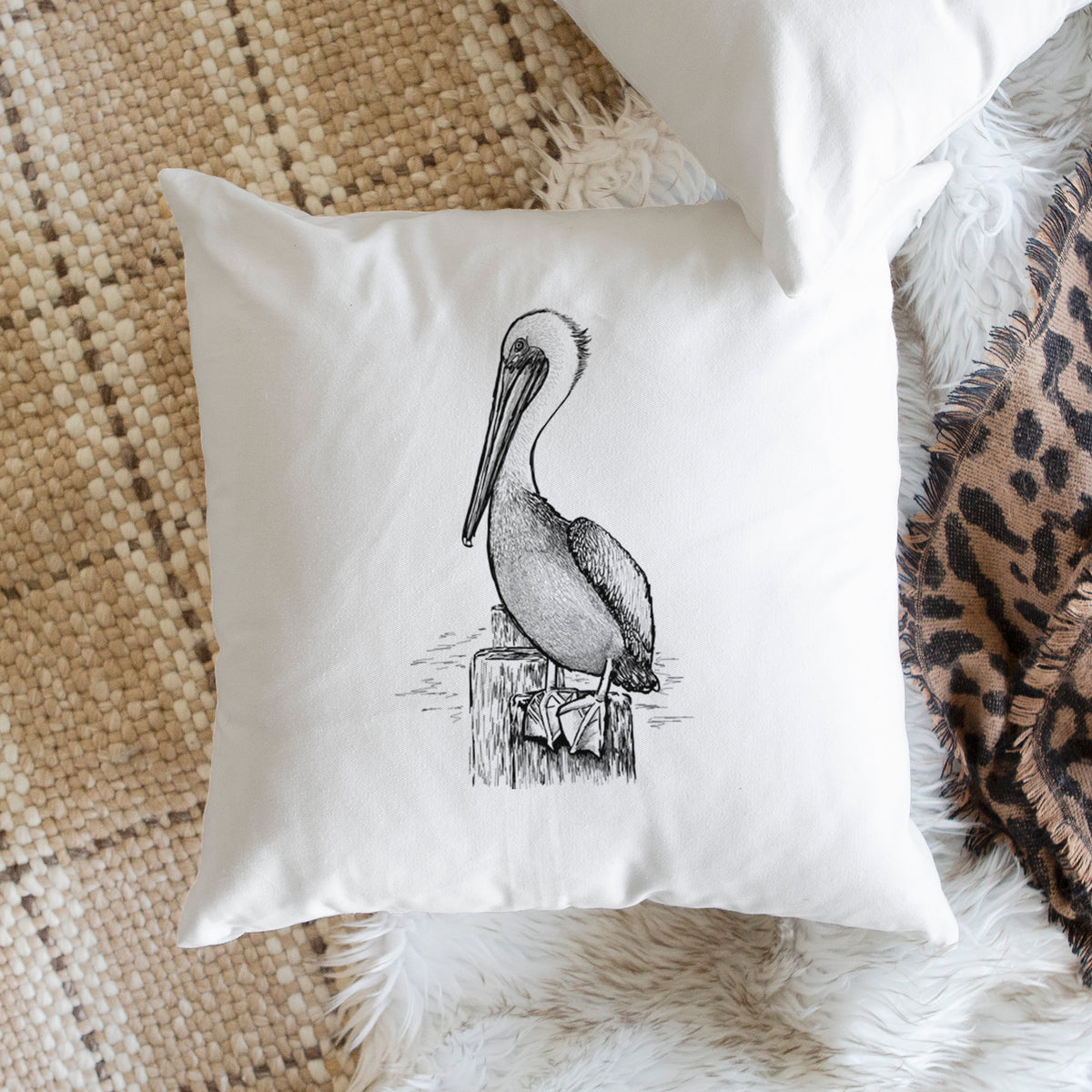 White pillow cover printed with a detailed black ink illustration of a brown pelican perched on a wooden piling, on a woven chair.
