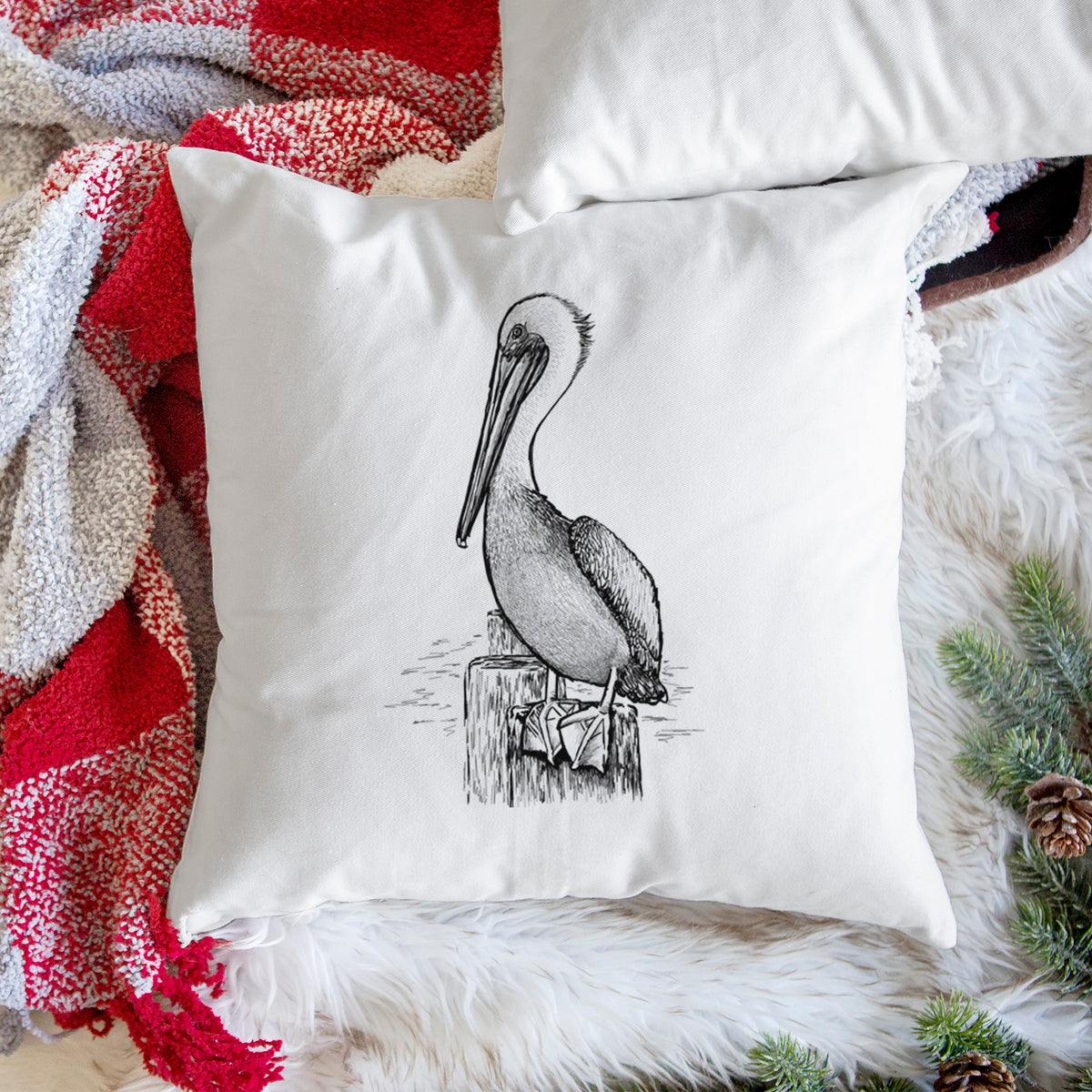 White pillow cover printed with a detailed black ink illustration of a brown pelican perched on a wooden piling, on a woven chair.