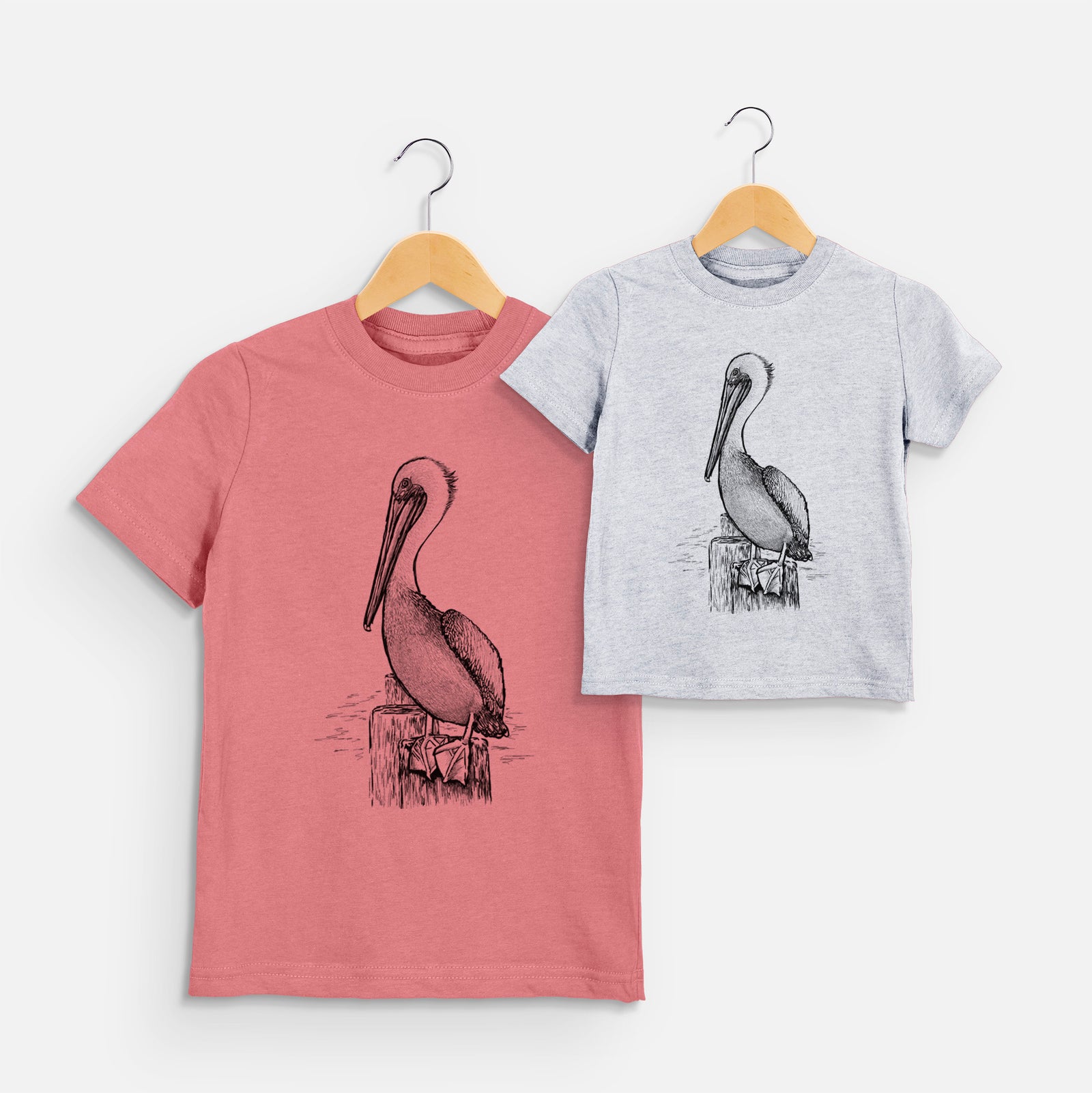 Two youth t-shirts in different colors on hangers, printed with a detailed black ink illustration of a brown pelican perched on a wooden piling.