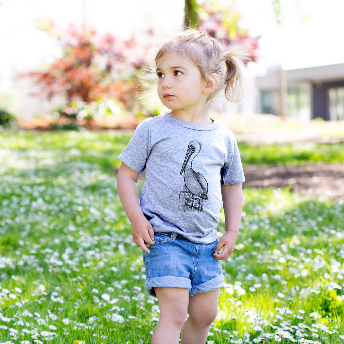 Toddler girl walking outdoors wearing a t-shirt printed with a detailed black ink illustration of a brown pelican perched on a wooden piling.