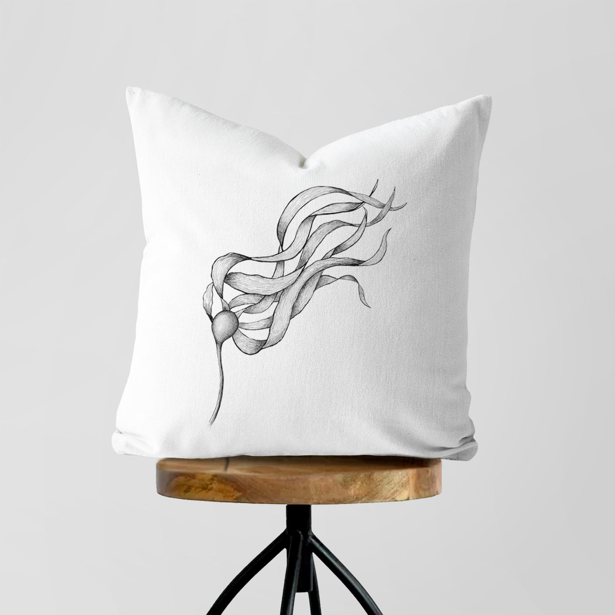 White pillow cover printed with a black ink line drawing of a strand of bull kelp with long flowing blades, on a wooden stool.
