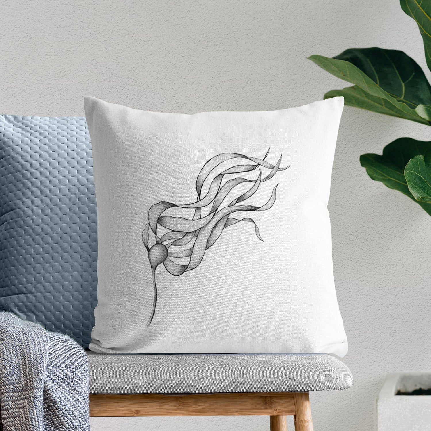White pillow cover printed with a black ink line drawing of a strand of bull kelp with long flowing blades, on a bench beside plants.