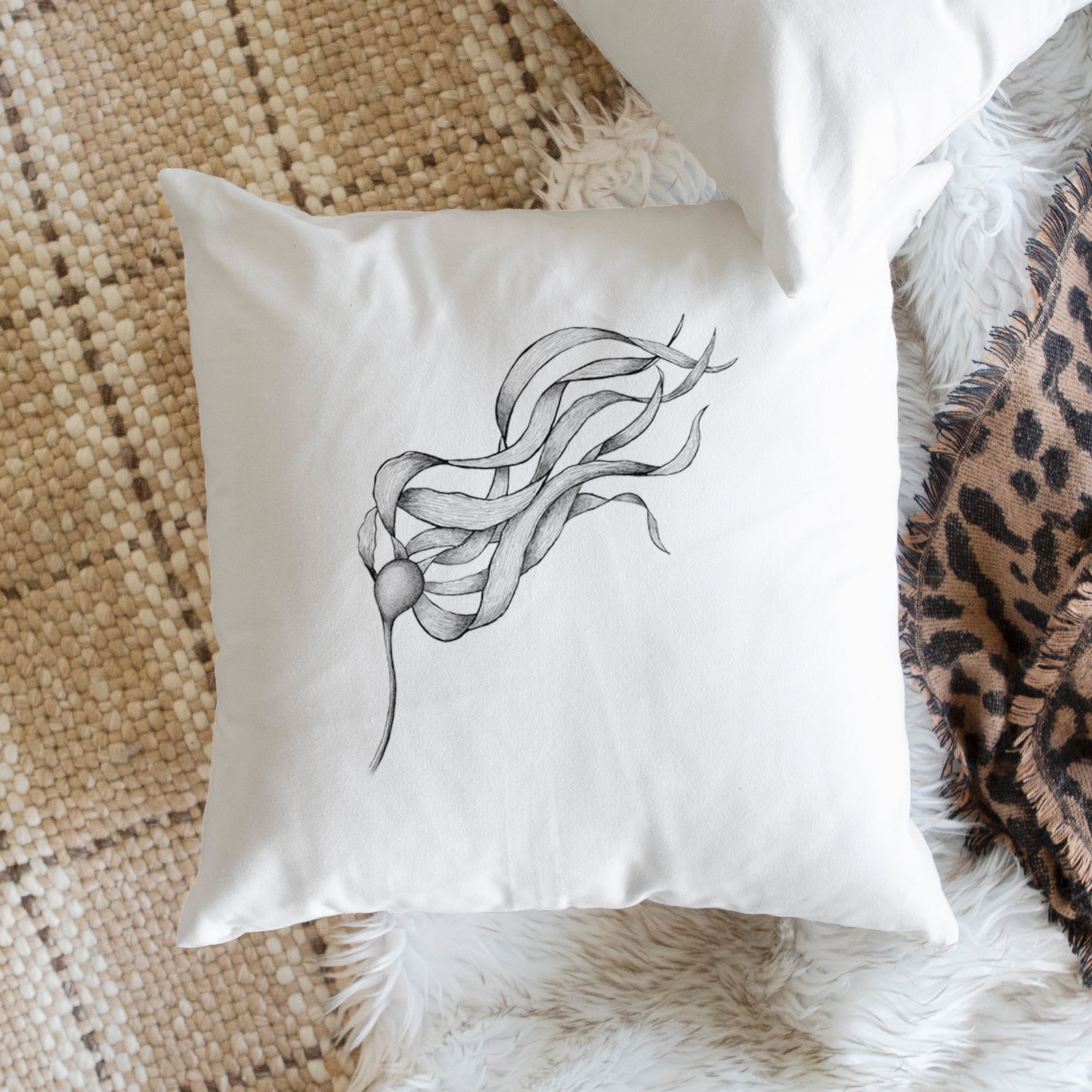 White pillow cover printed with a black ink line drawing of a strand of bull kelp with long flowing blades, on a bench beside plants.