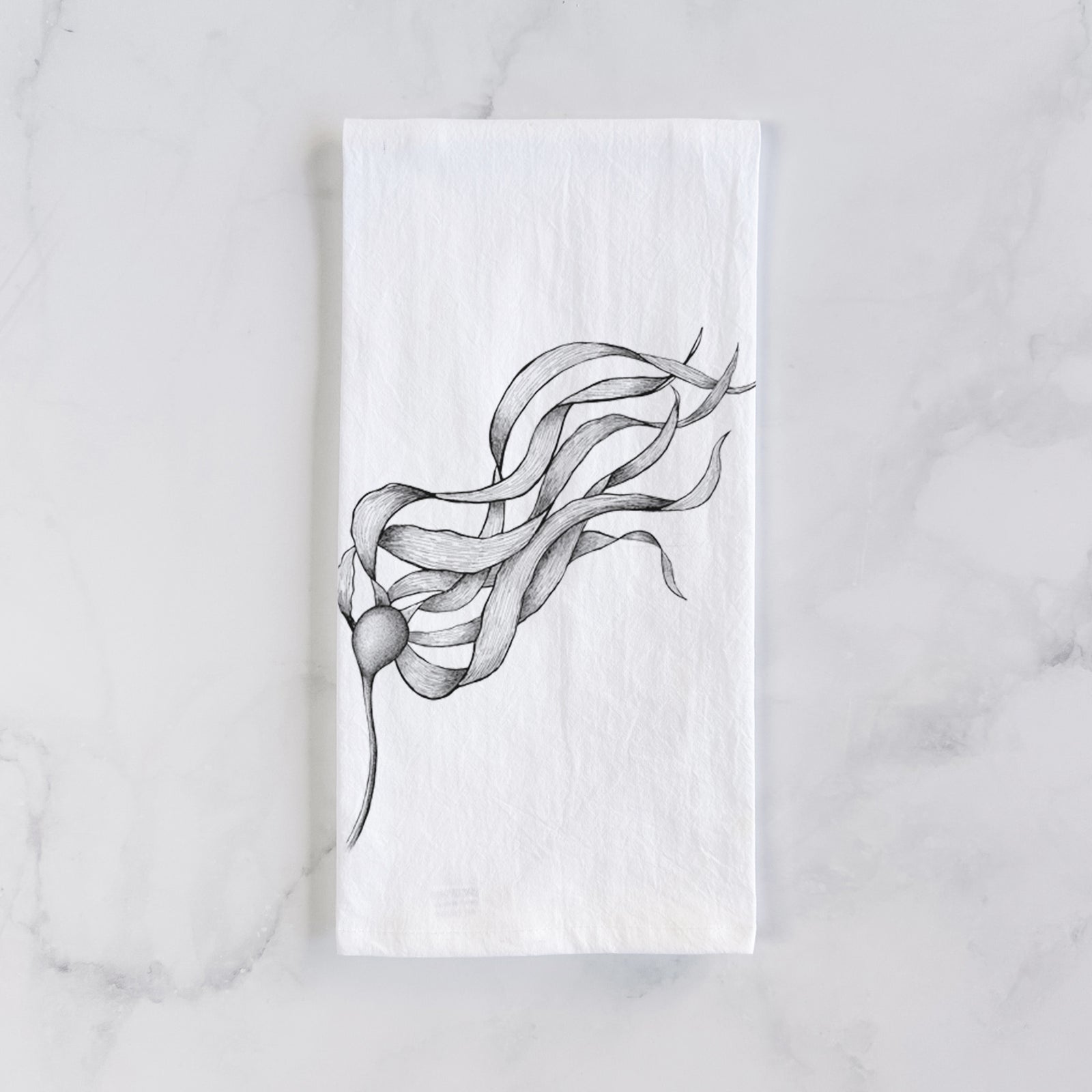 White cotton tea towel printed with a black ink line drawing of a strand of bull kelp with long flowing blades, on a marble counter beside a round wooden cutting board and greenery.