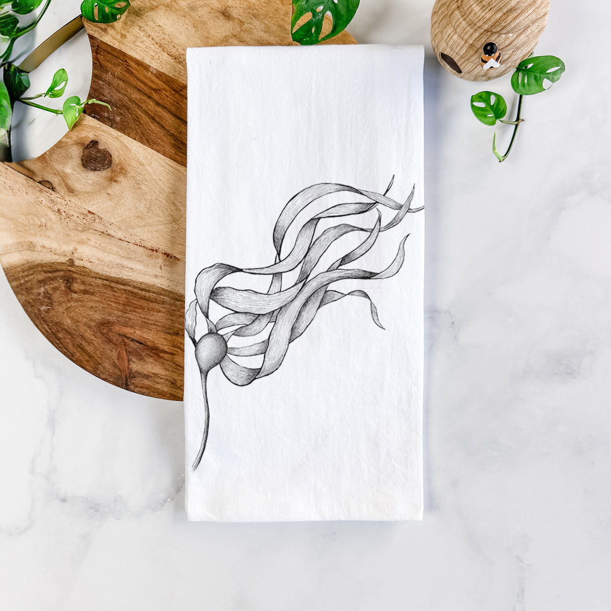 White cotton tea towel printed with a black ink line drawing of a strand of bull kelp with long flowing blades, on a marble counter beside a round wooden cutting board and greenery.