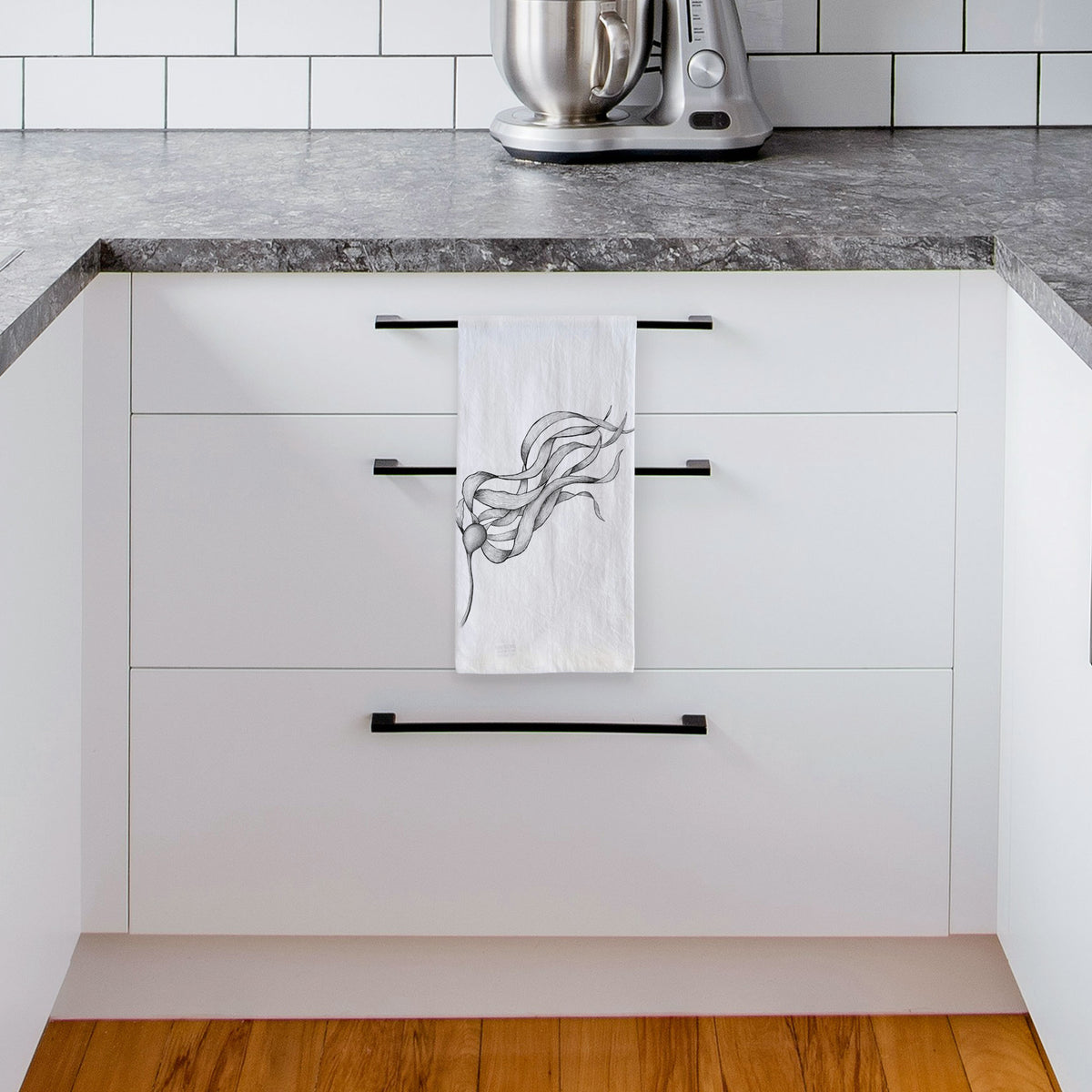 White tea towel printed with a black ink line drawing of a strand of bull kelp with long flowing blades, hanging from a drawer handle in a bright white kitchen.