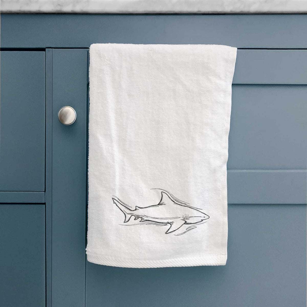 White hand towel printed with a black ink illustration of a bull shark swimming, hanging on a cabinet door.